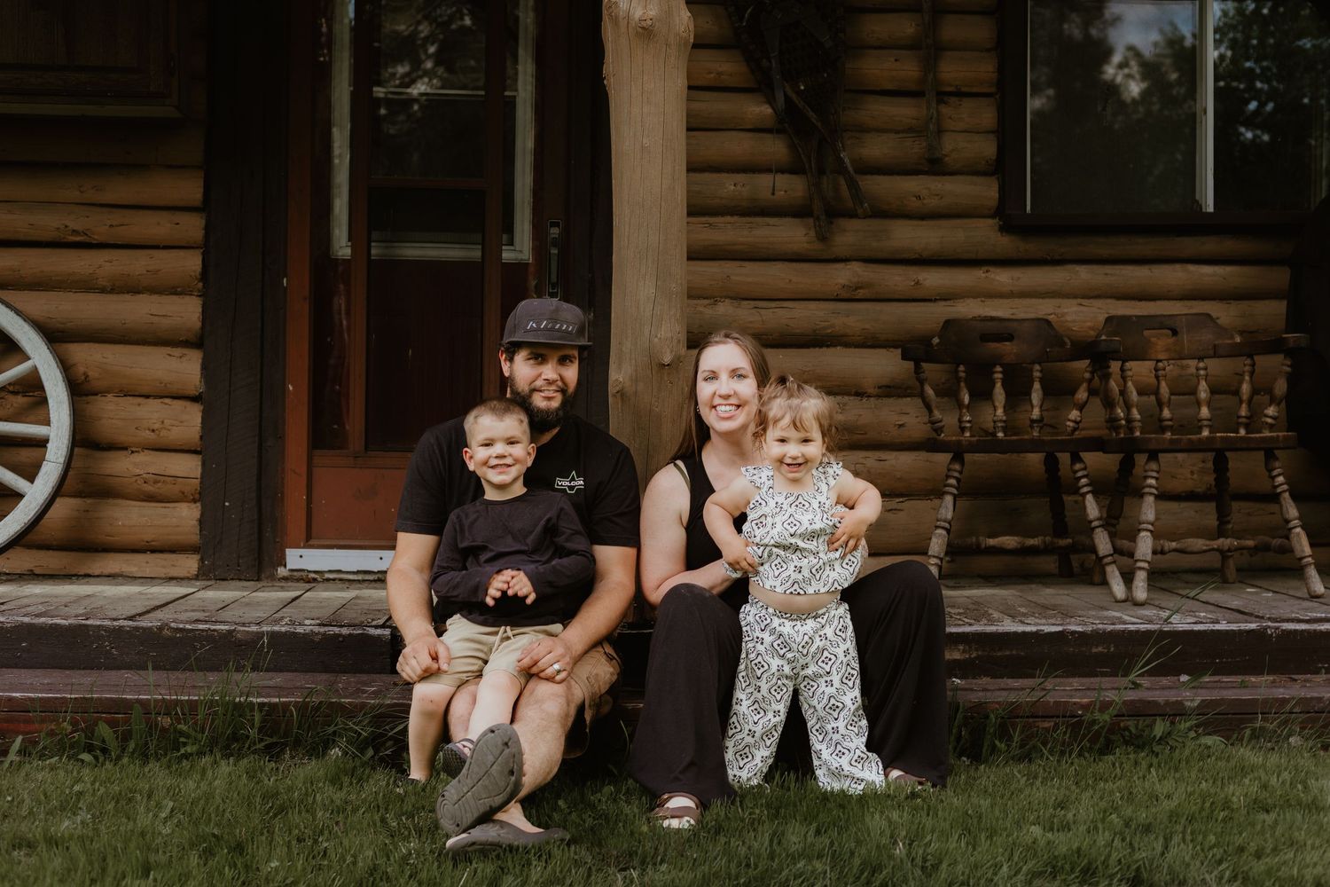 Family of four smiling on log cabin porch steps, parents holding young children outdoors.