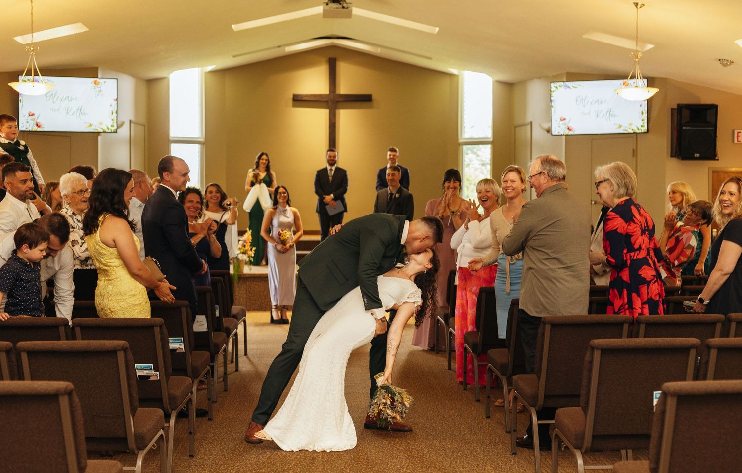 Groom dips bride for a kiss in church aisle during wedding ceremony, guests applauding, cross visible at altar.
