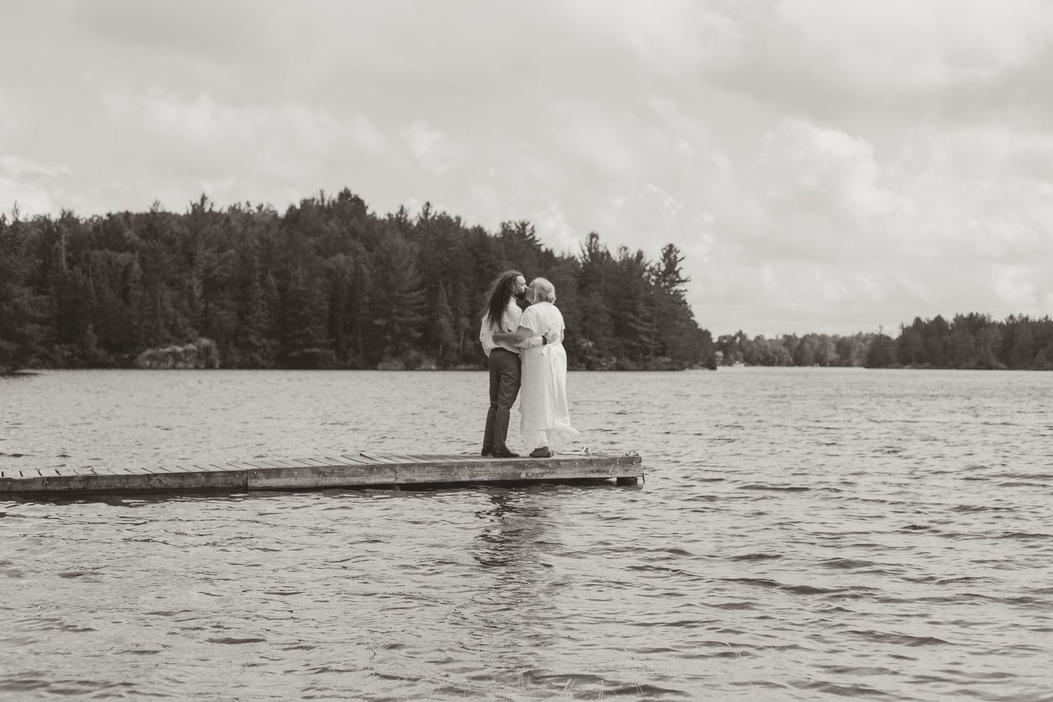 Black and white photo of newlyweds embracing on a wooden dock surrounded by a serene lake.