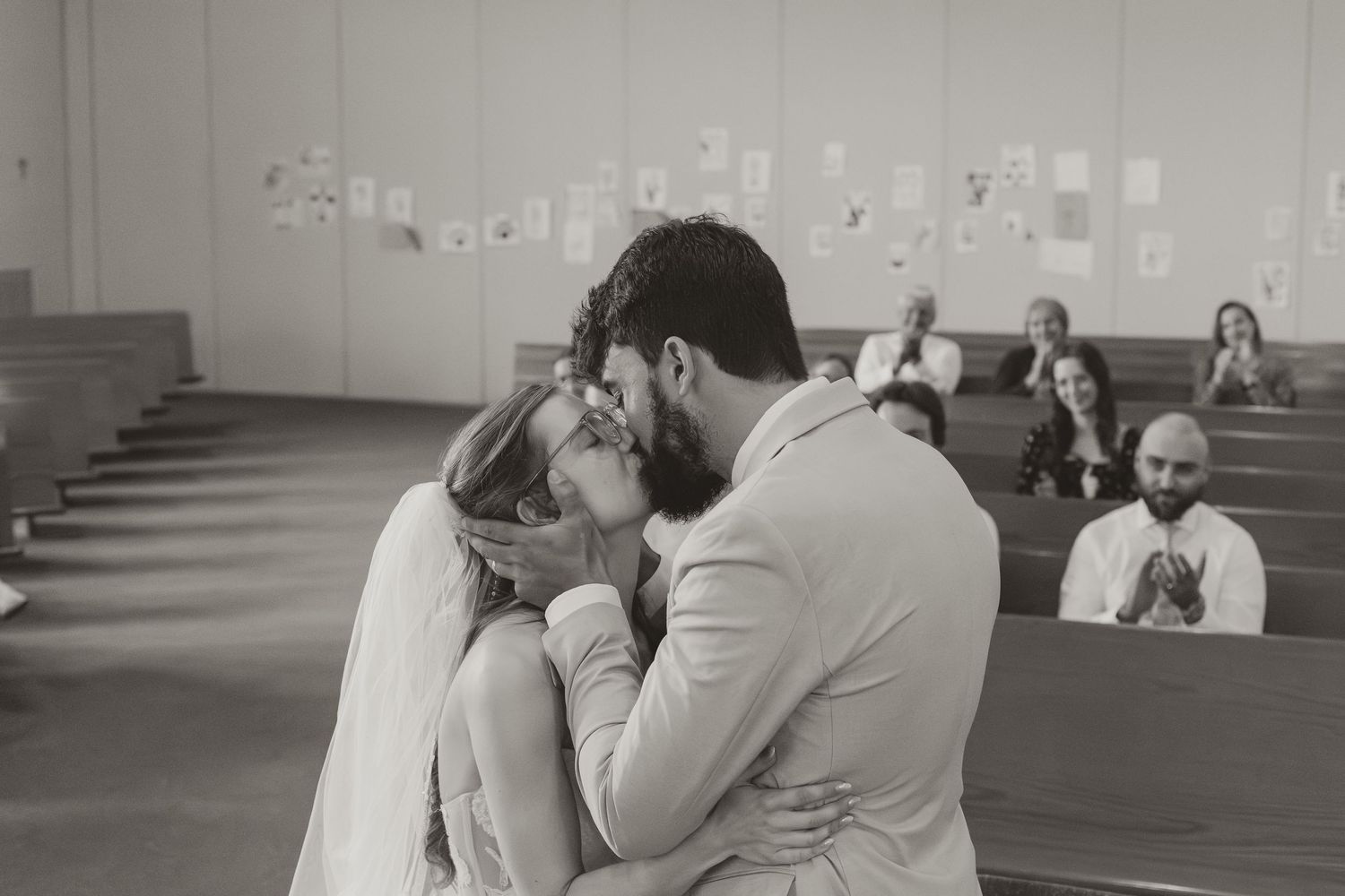 Black and white photo of newlyweds sharing first kiss at church altar while guests applaud in background.