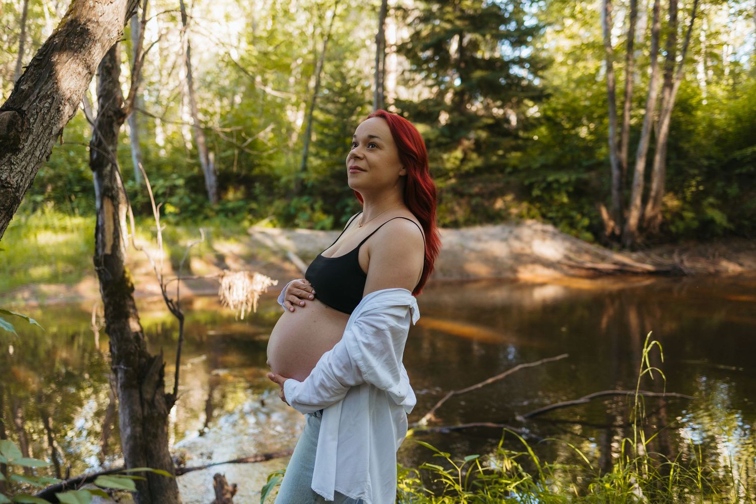 Pregnant woman with red hair cradling her baby bump outdoors near a sunlit forest pond on a warm day.