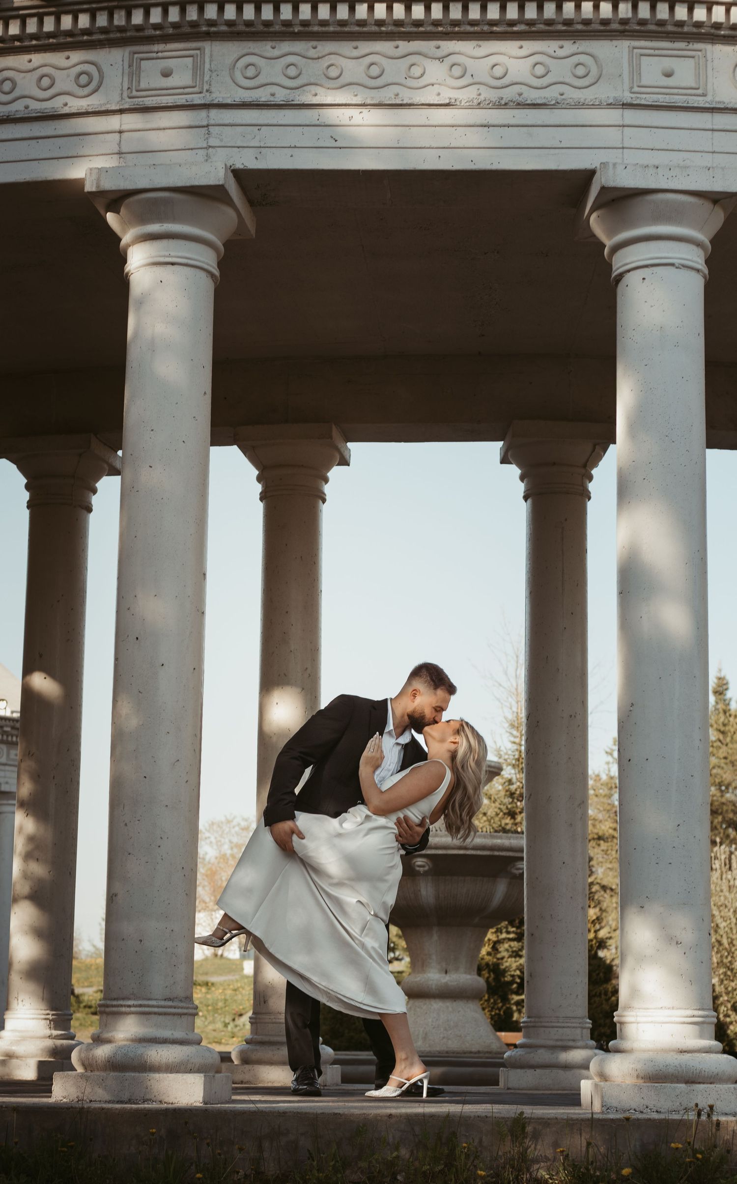 Couple sharing a romantic dip kiss under a classical stone colonnade with elegant white columns.