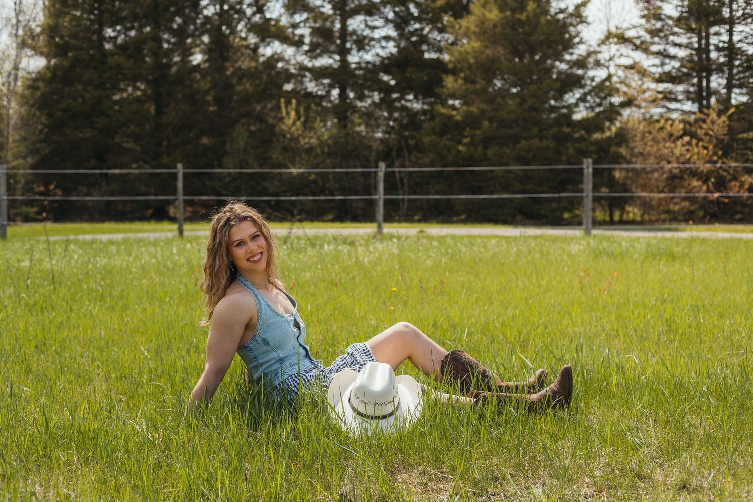 Young woman in denim romper and cowboy boots sitting in green field with white hat, smiling outdoors.