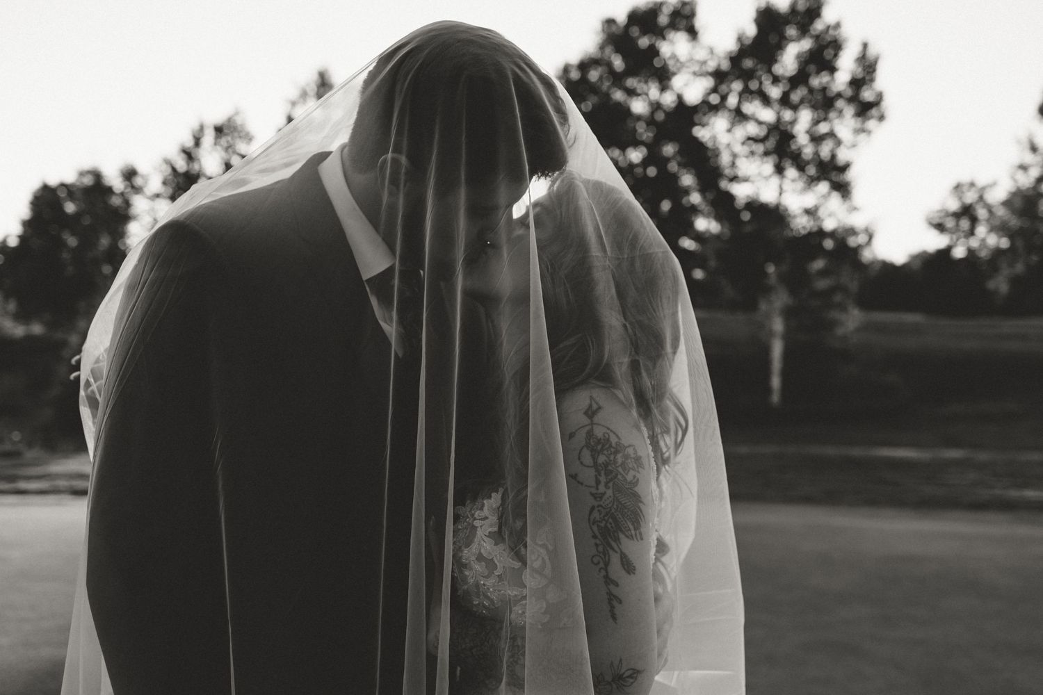 Black and white photo of bride and groom sharing a kiss under a wedding veil outdoors.