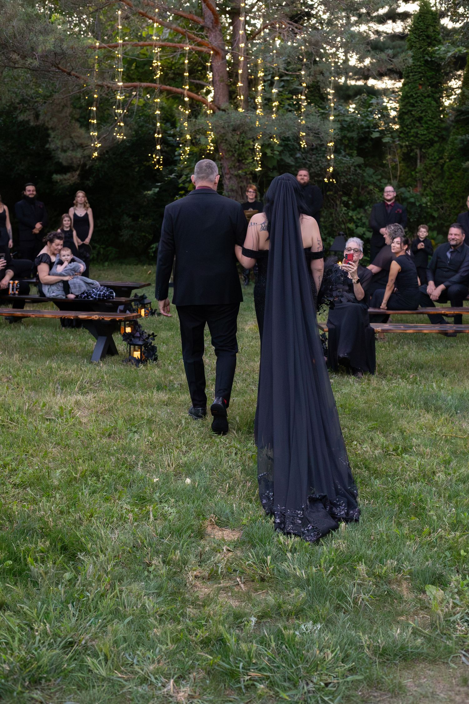 Gothic wedding ceremony outdoors with bride in black veil and groom walking toward altar among guests.