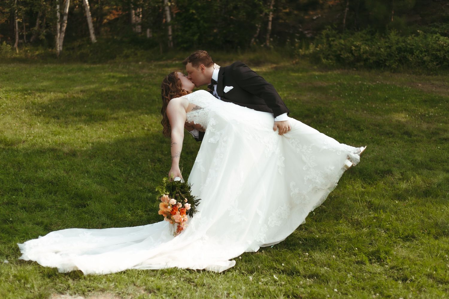 Groom dipping bride in white gown while holding orange bouquet on green lawn.