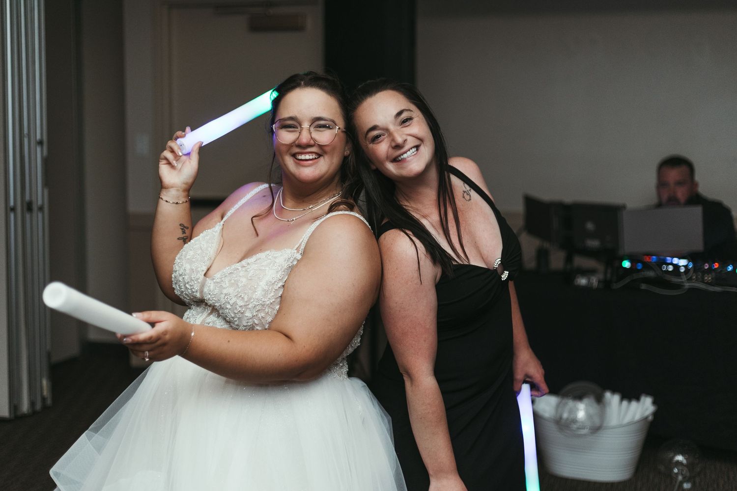 Bride in white gown and bridesmaid in black dress holding colorful glow sticks at wedding reception.