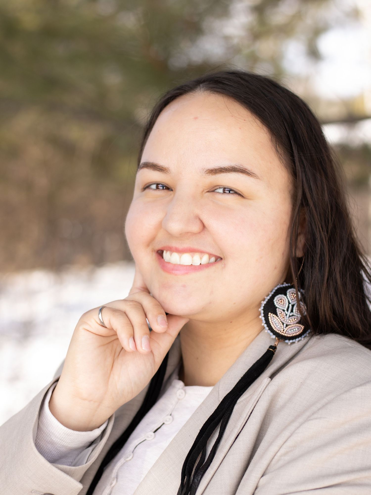 A smiling young woman with dark hair wearing beaded earrings and a beige blazer poses outdoors in winter.