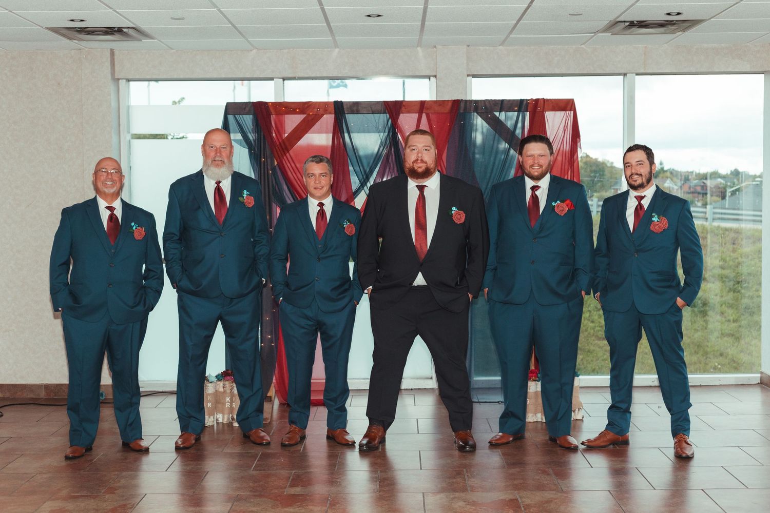 Six groomsmen in navy suits with red boutonnieres posing together in front of colorful draped backdrop.
