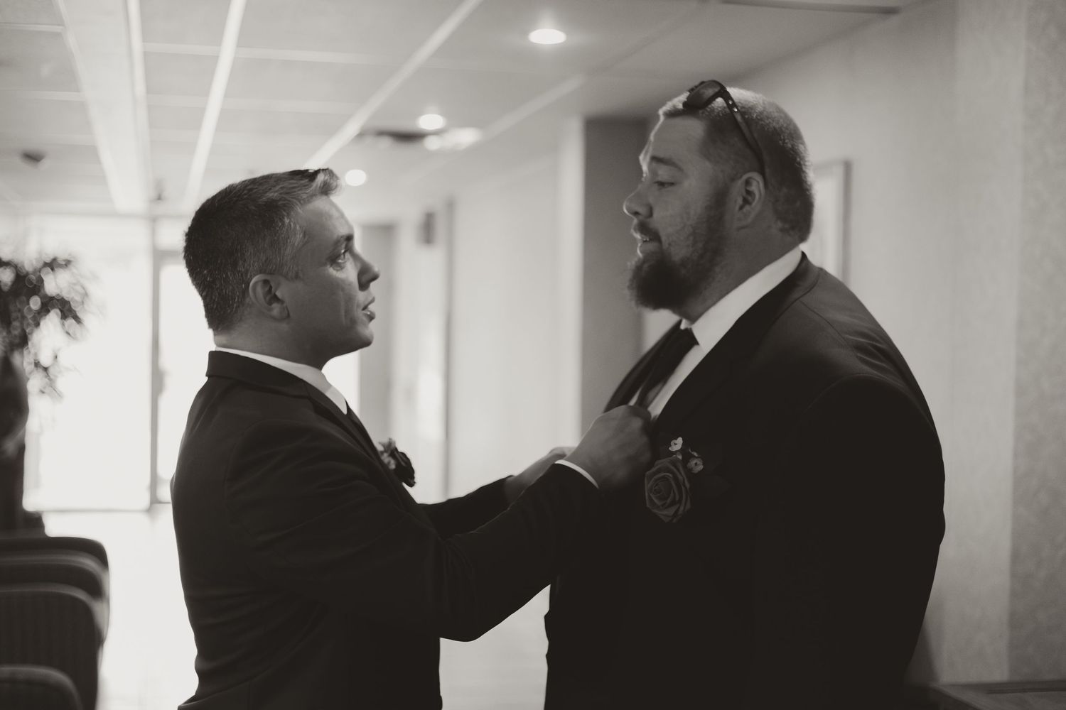 Black and white photo of two men in suits adjusting a tie before a wedding ceremony.