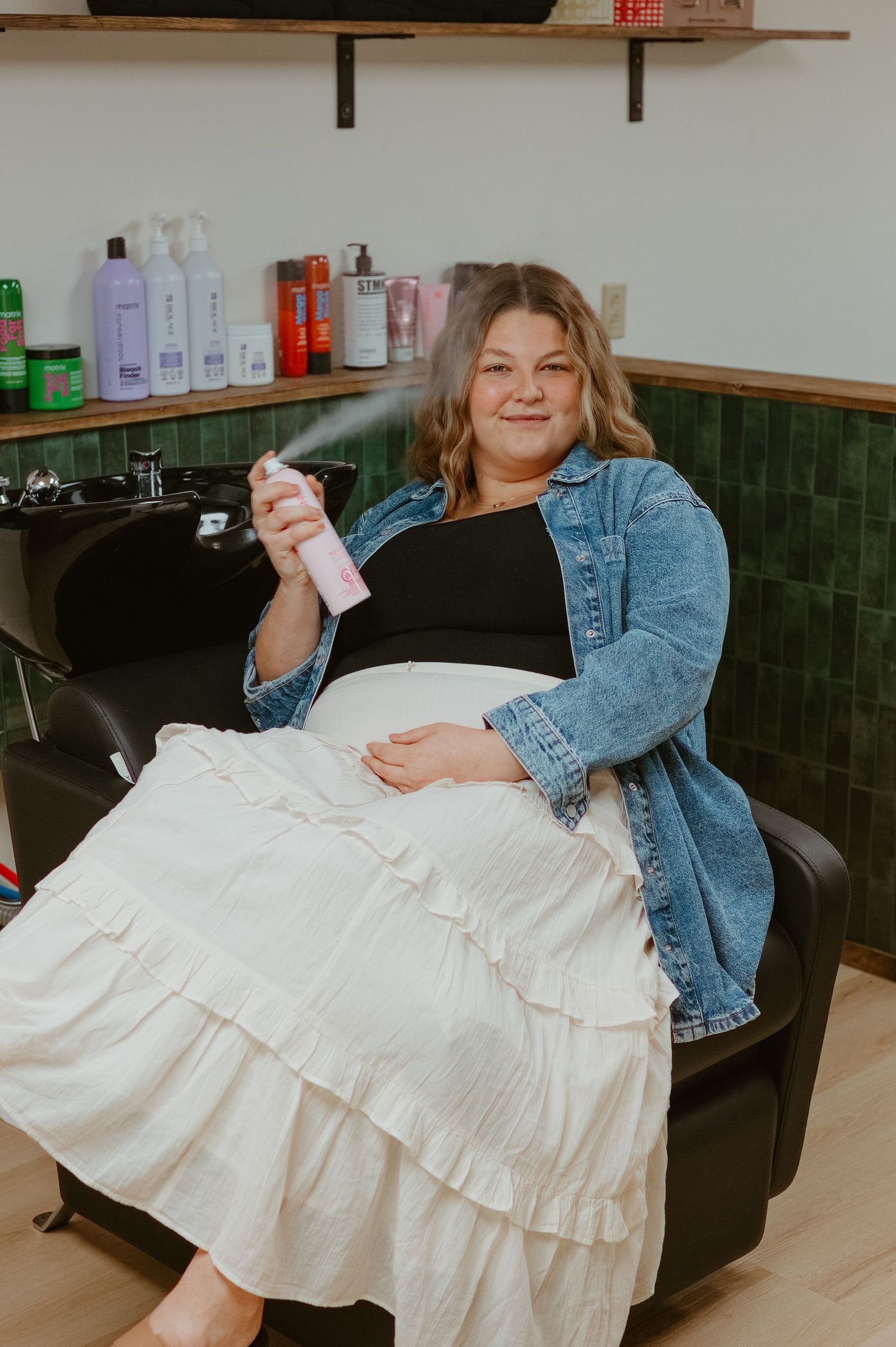 Woman sitting in salon chair smiling, wearing denim jacket and white skirt, spraying hair product near shampoo bowls.