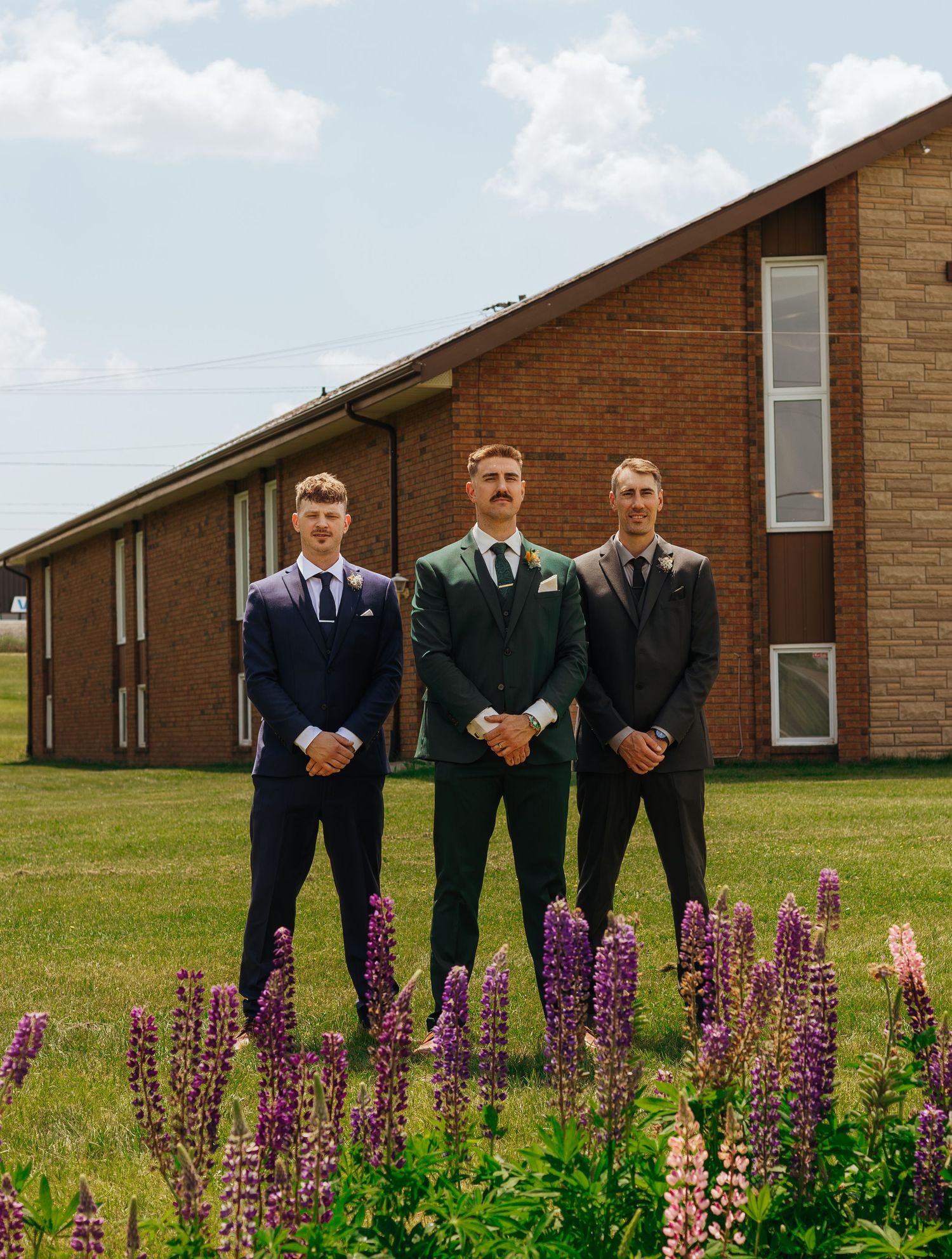 Three groomsmen in suits stand before a brick church surrounded by blooming purple and pink lupine flowers.
