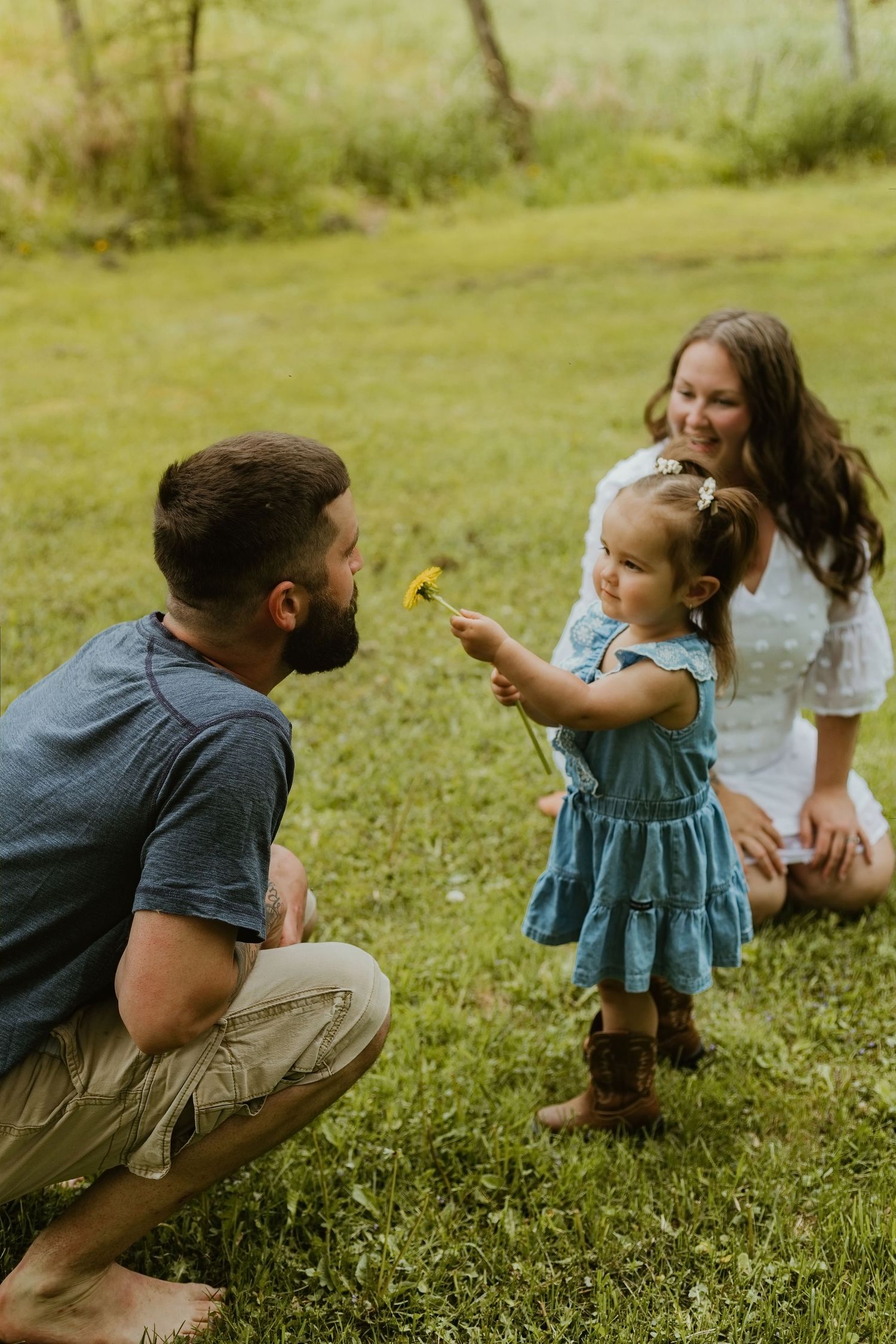A toddler girl in a blue dress offers a dandelion to her bearded father while mom watches and smiles outdoors.