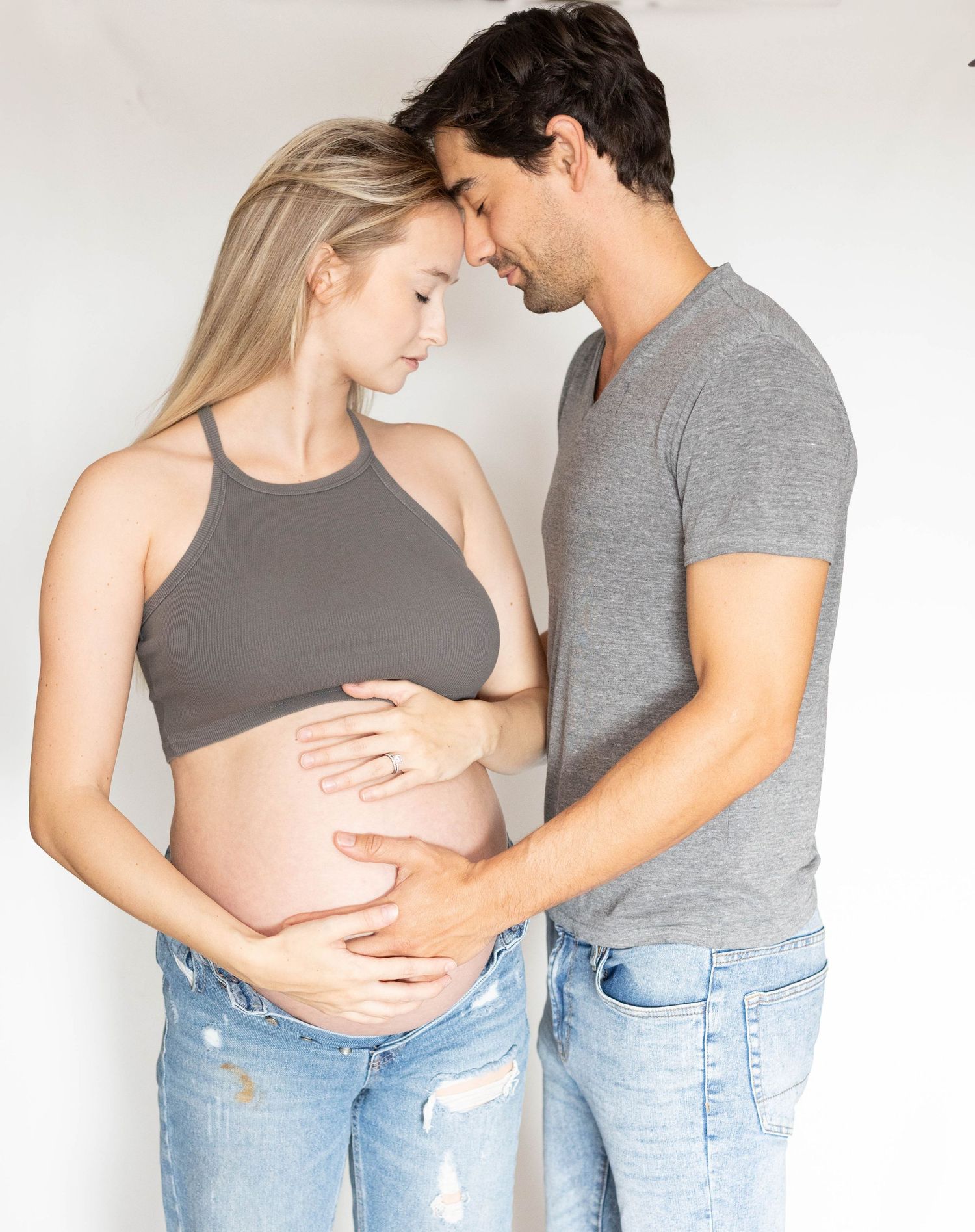Expecting couple tenderly holding pregnant belly together in matching gray and jeans outfits indoors.