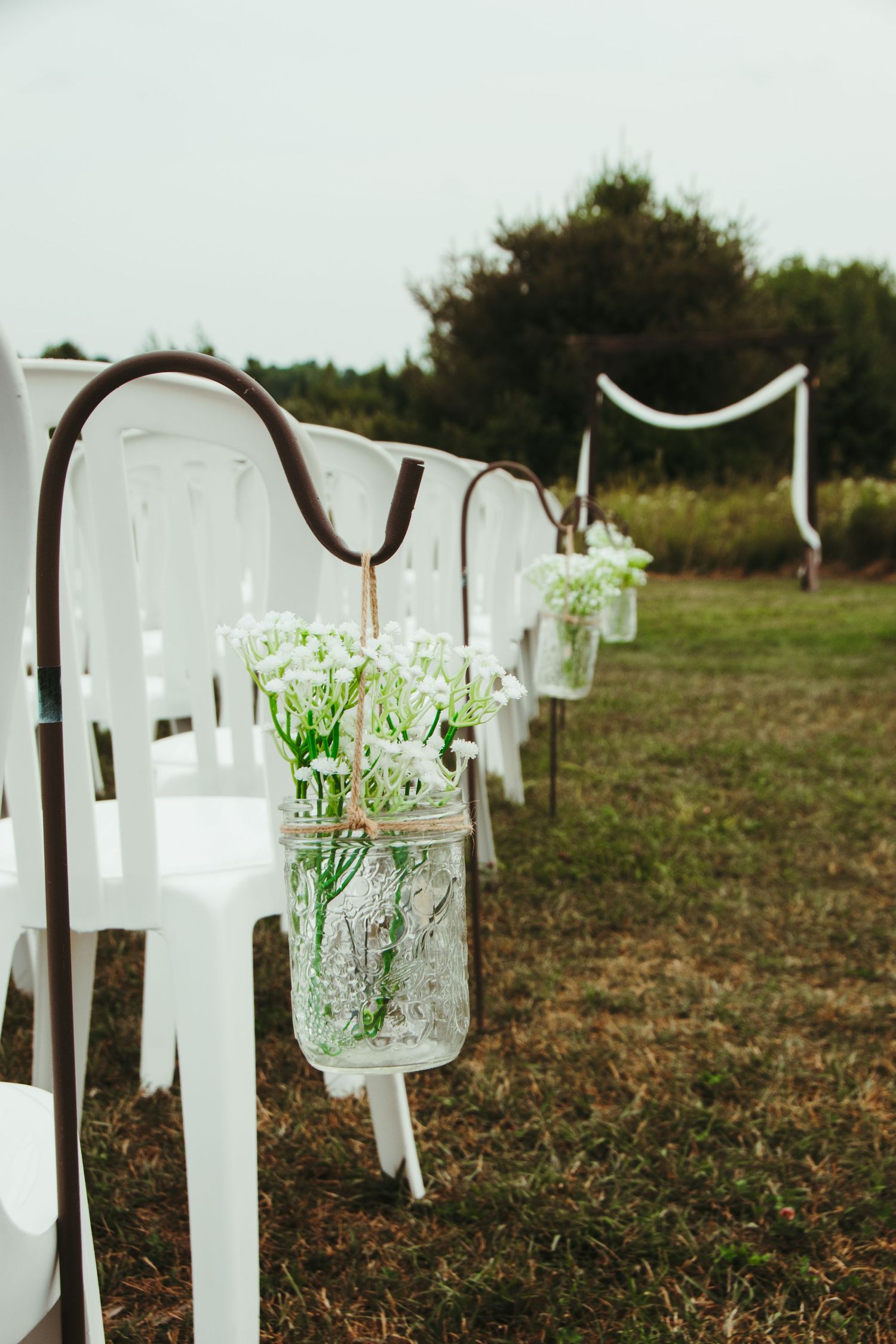 Mason jar floral arrangements hanging from shepherd hooks along outdoor wedding aisle.