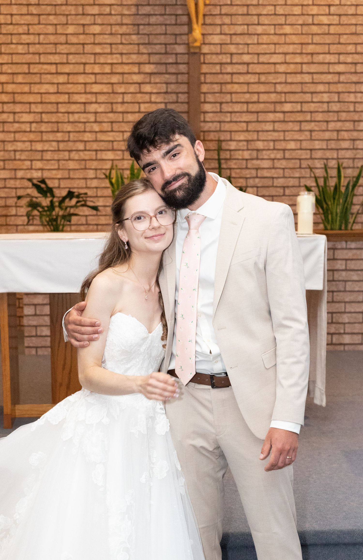 Bride and groom pose together smiling in a church, she in a white lace gown and he in a beige suit with pink tie.