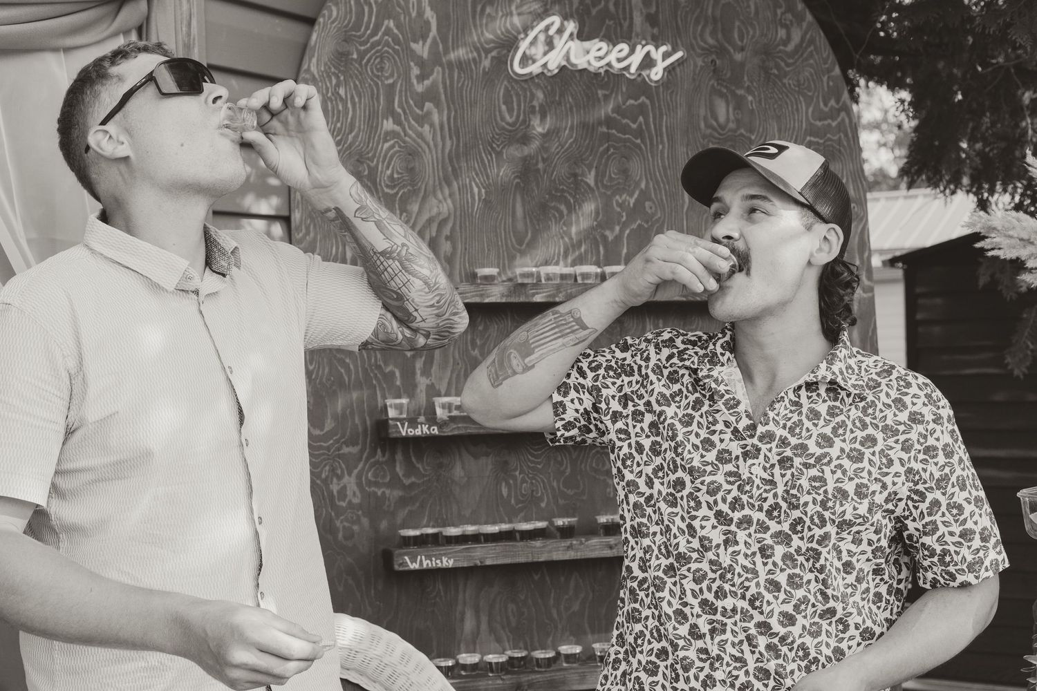 Two men taking shots in front of a Cheers sign at an outdoor wedding celebration.