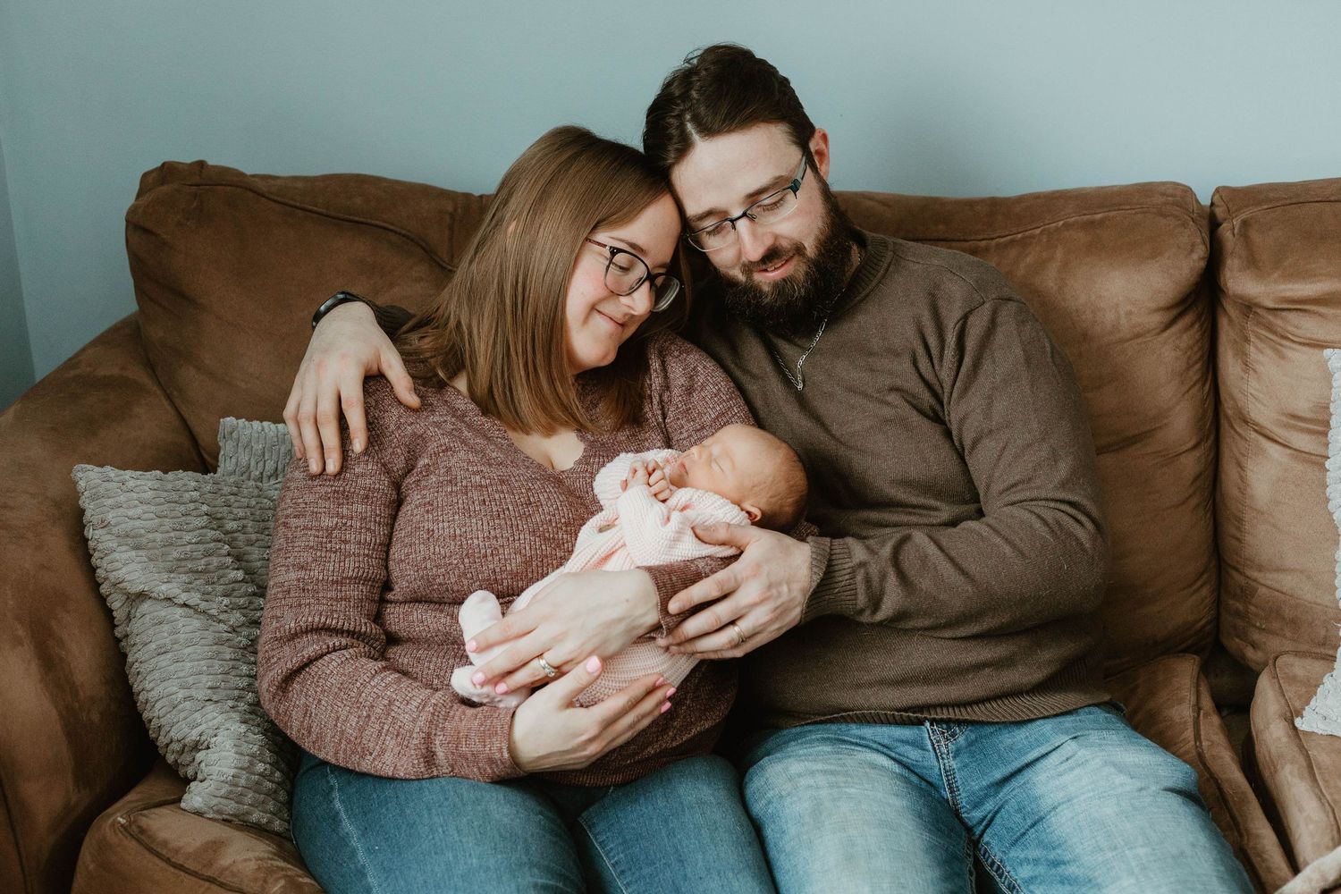 New parents sitting on a brown leather sofa smiling while cradling their newborn baby.