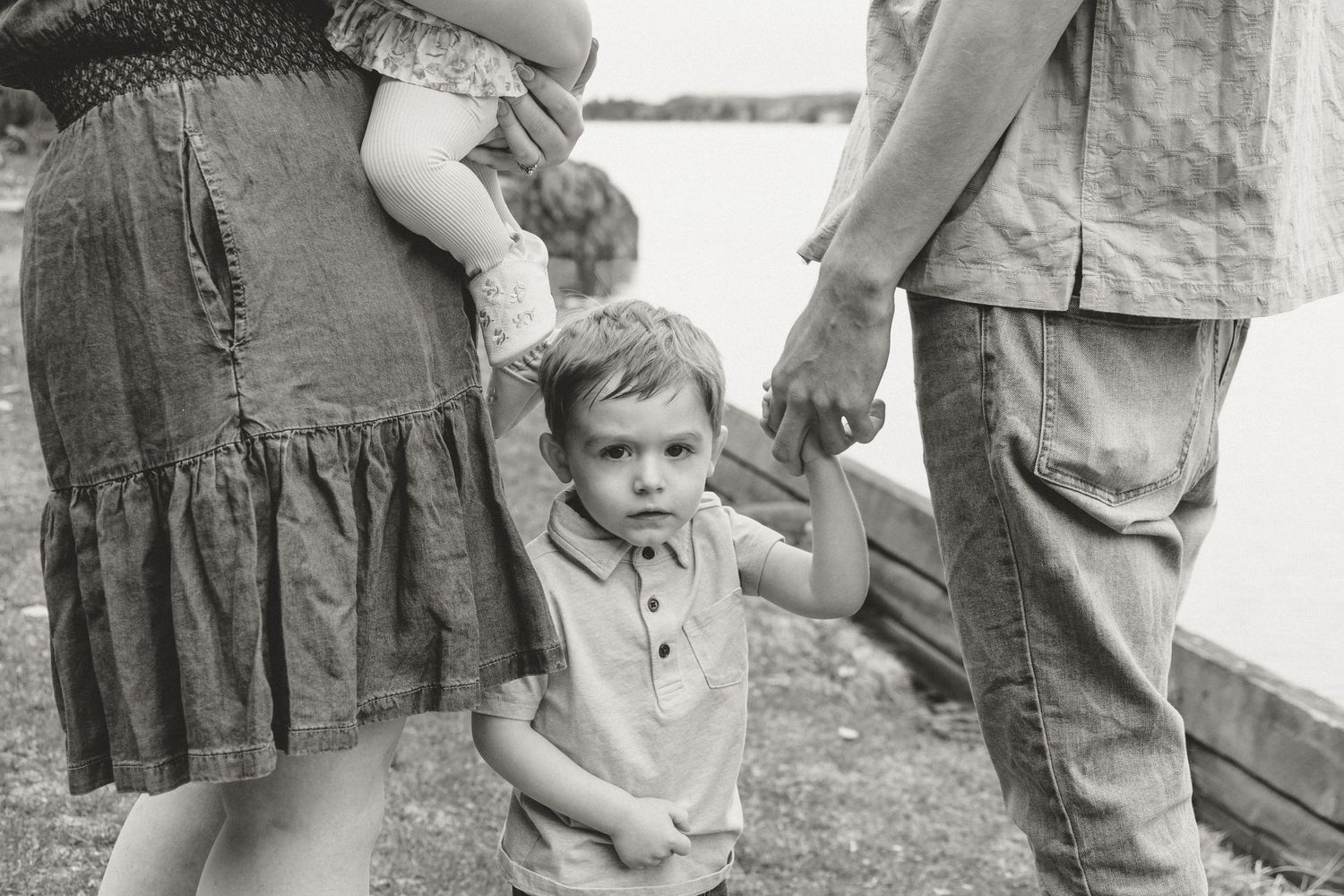 Young toddler boy holding parents' hands outdoors near a lake in black and white photo.
