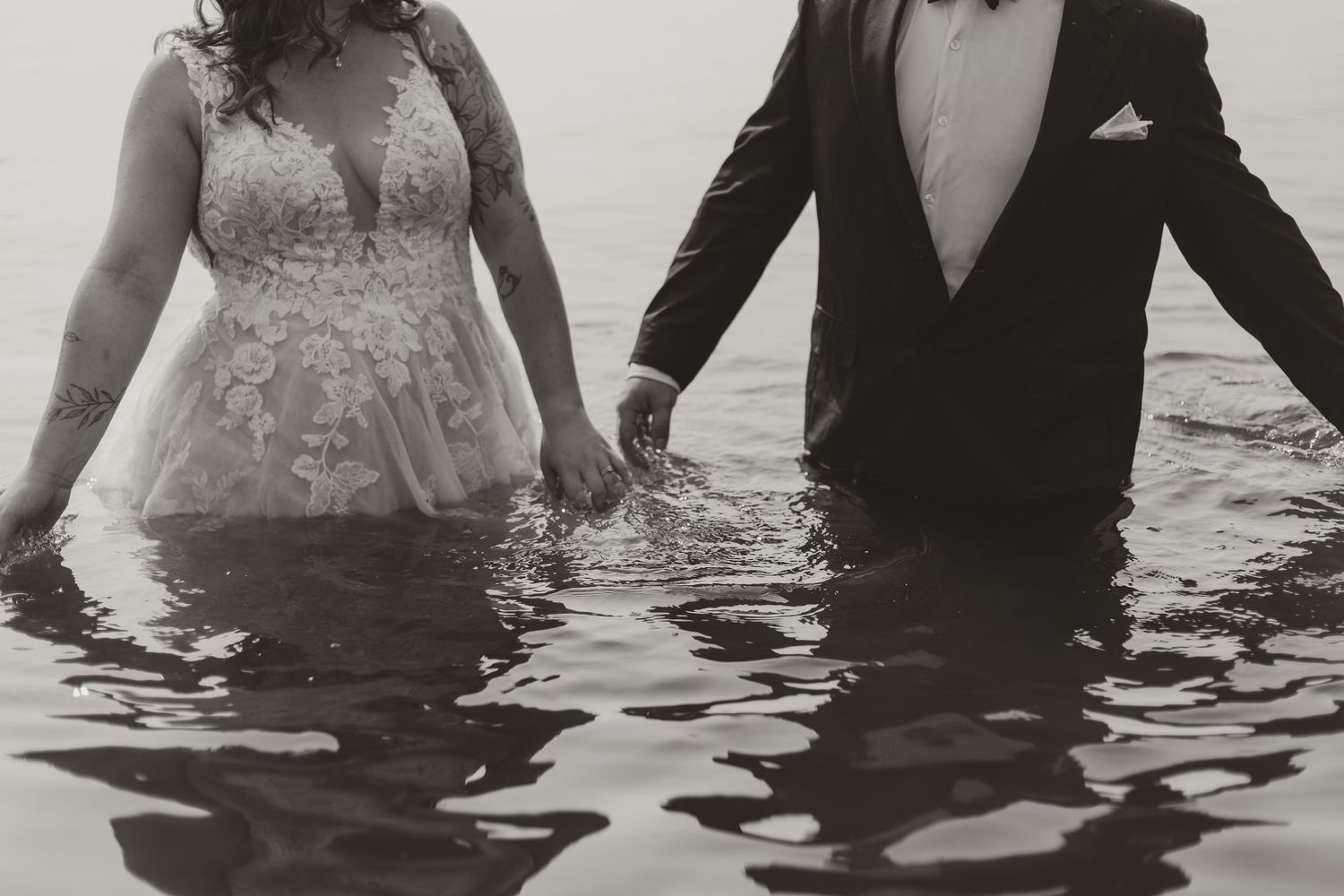 Black and white photo of bride and groom holding hands while standing in water during wedding shoot.