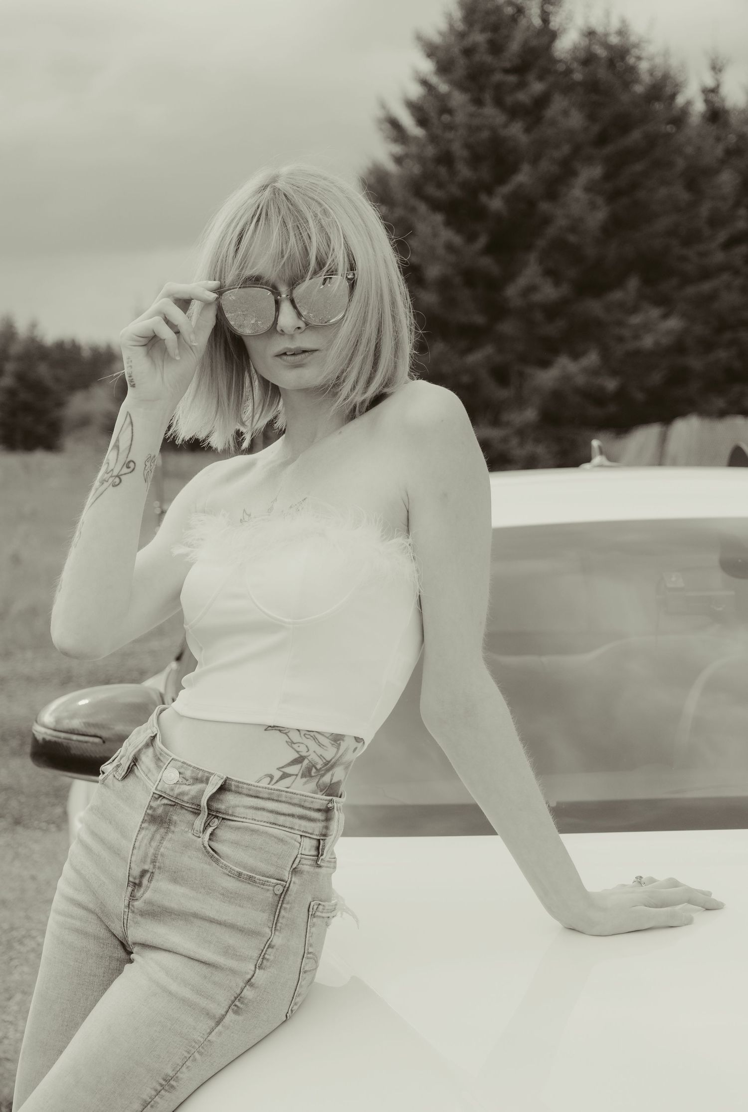 Stylish blonde woman in white crop top and jeans poses beside white car in black and white photo.