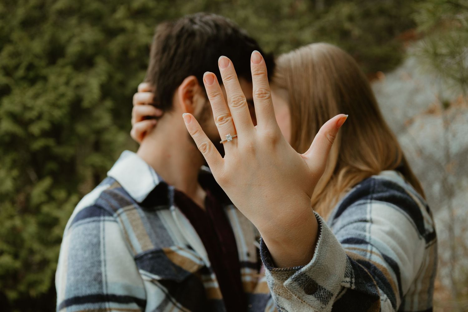 Couple kisses outdoors as woman displays engagement ring proudly to the camera.