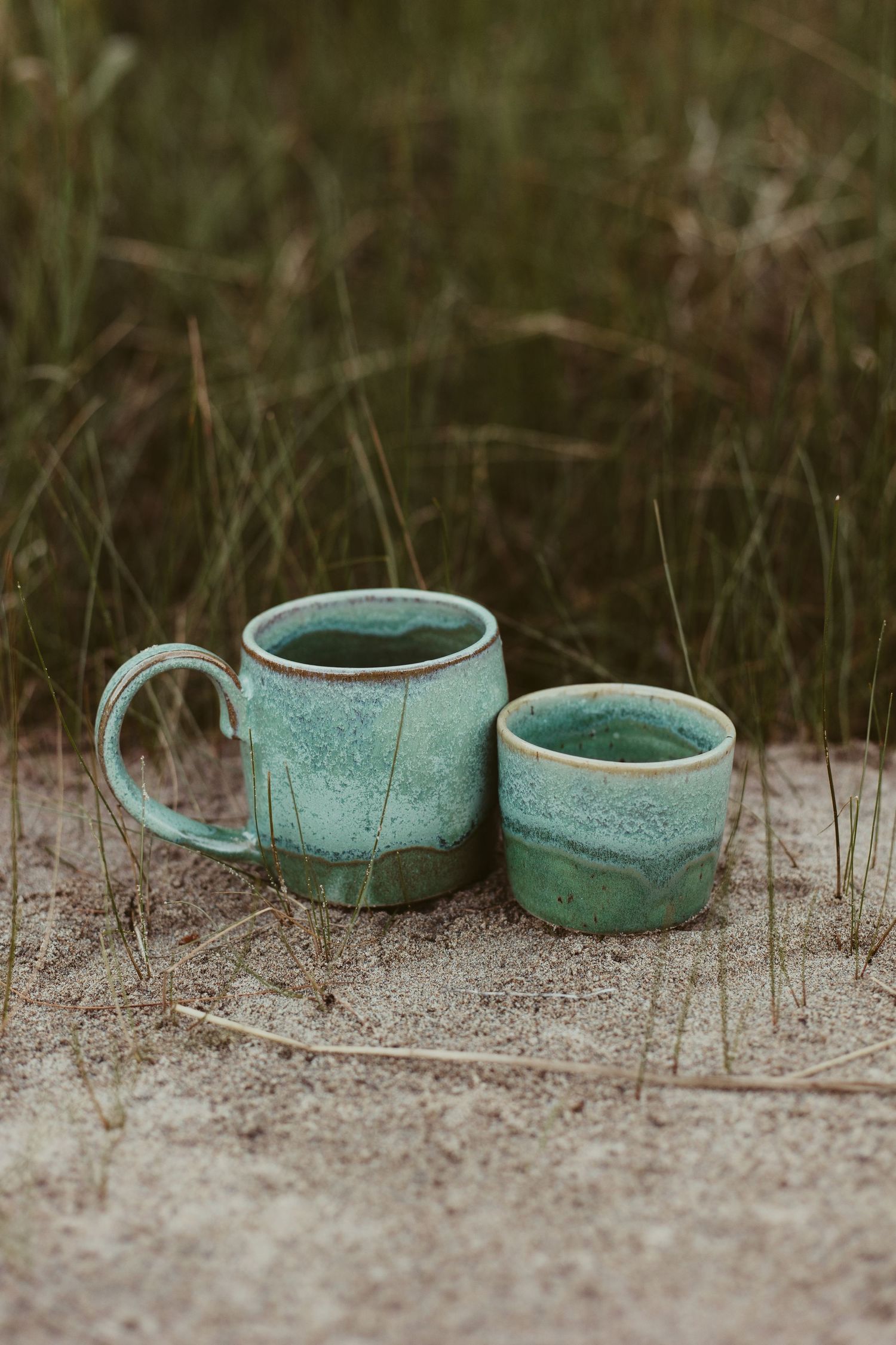 Two rustic teal glazed ceramic mugs sitting on sandy ground with green grass background.