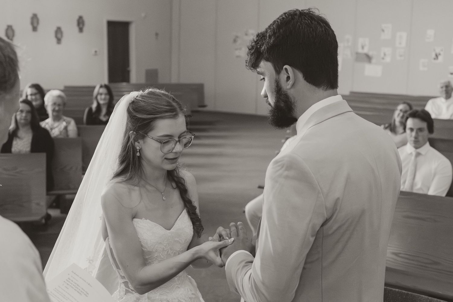 Bride placing wedding ring on grooms finger during church ceremony with guests seated in pews behind.