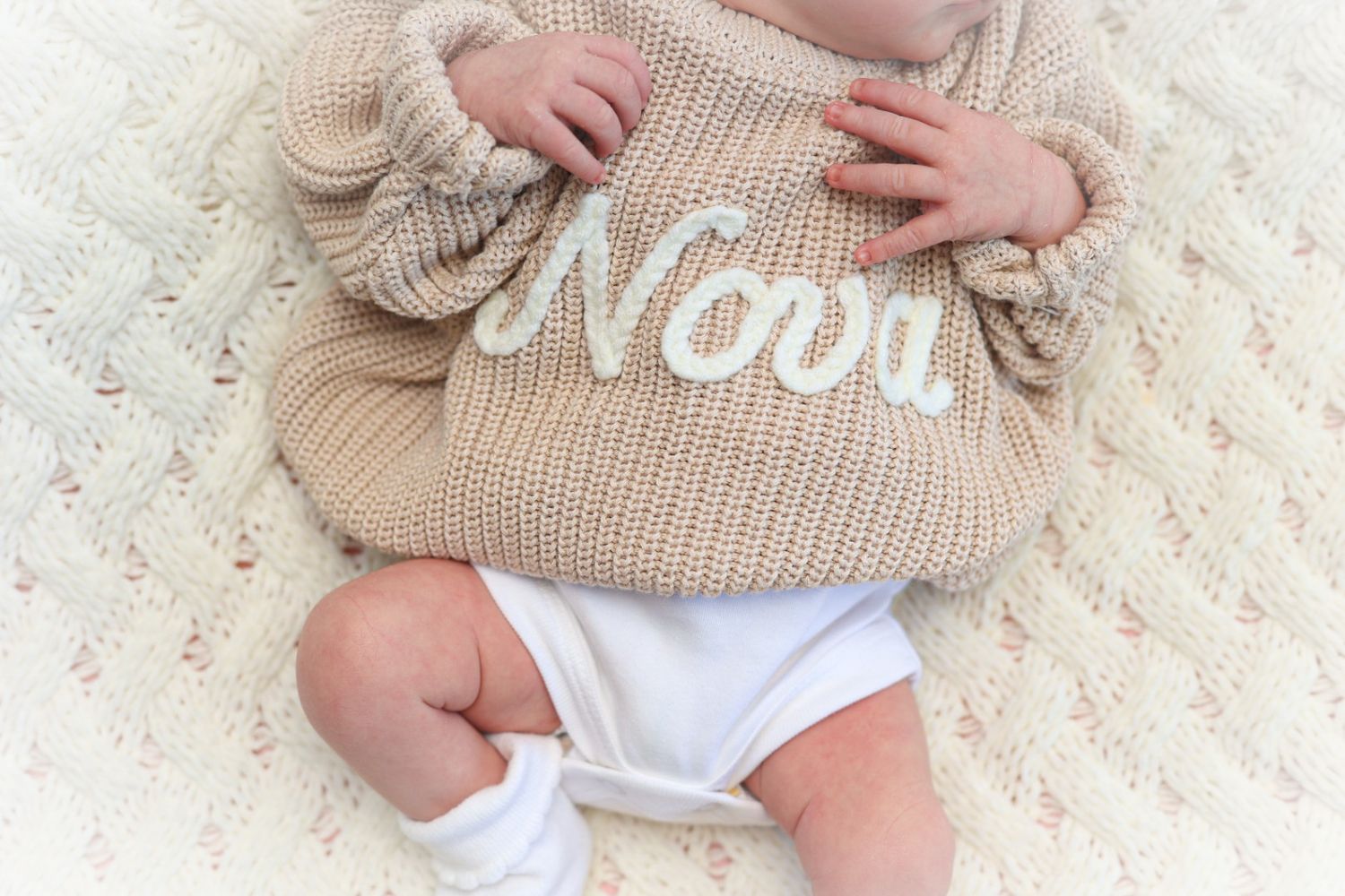 A newborn baby wearing a personalized beige knit sweater with the name Nova on a cream blanket.