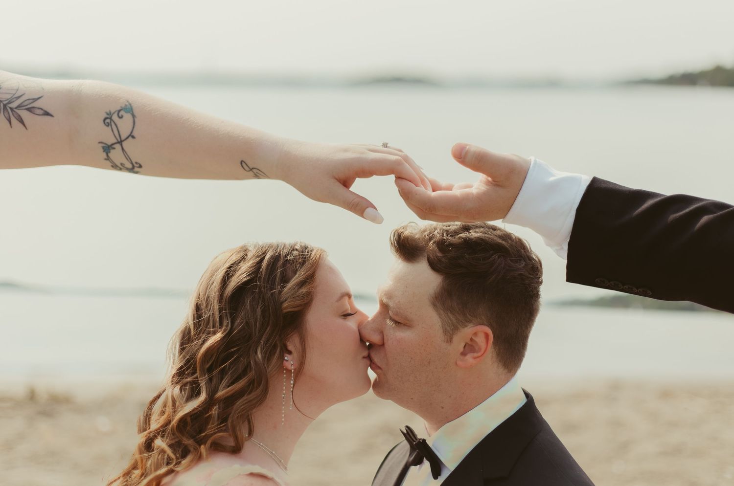 Bride and groom share a romantic beach kiss while hands form a heart above them.