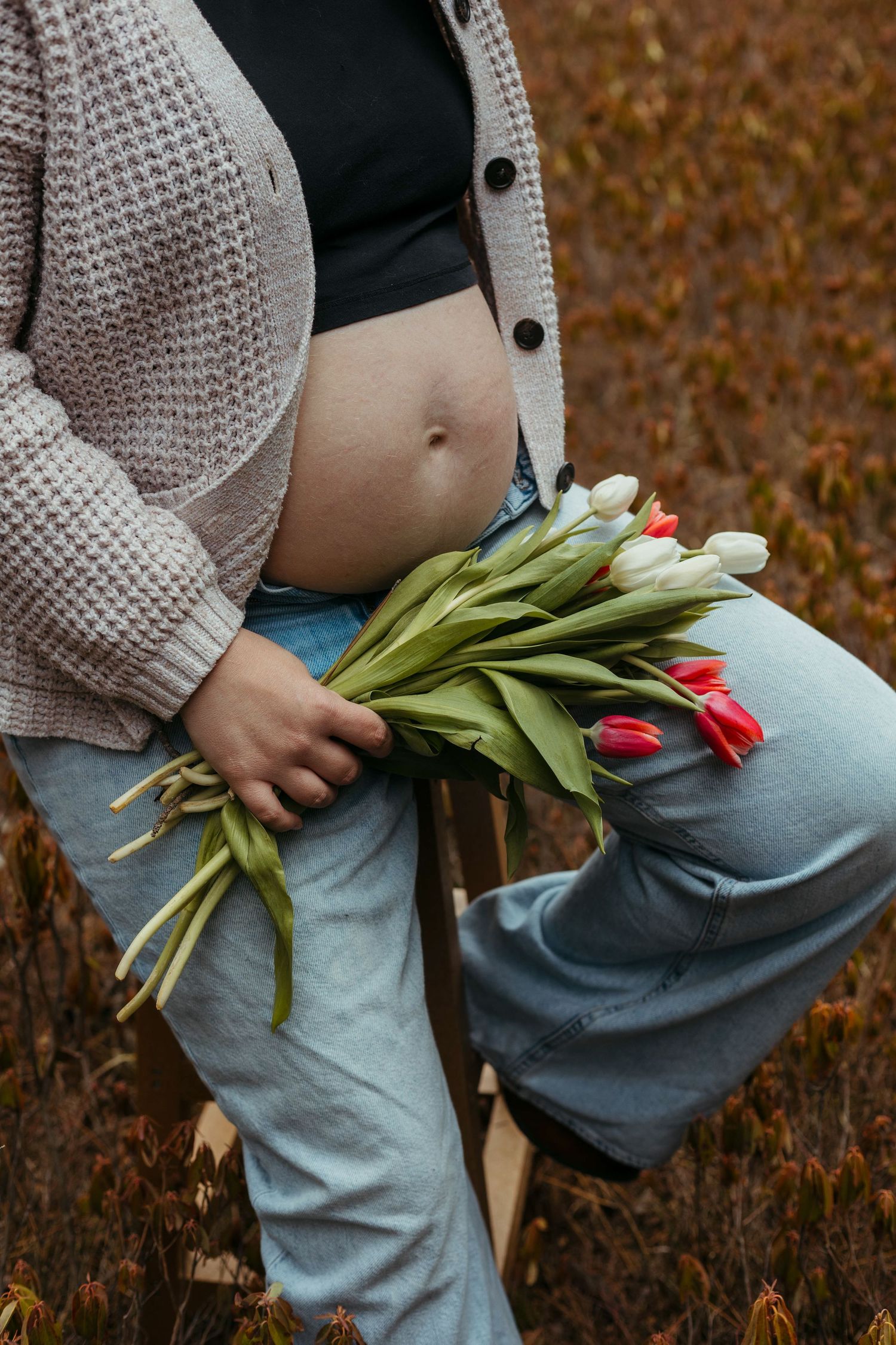 A pregnant woman holds colorful tulips against her bare belly in an autumn field.