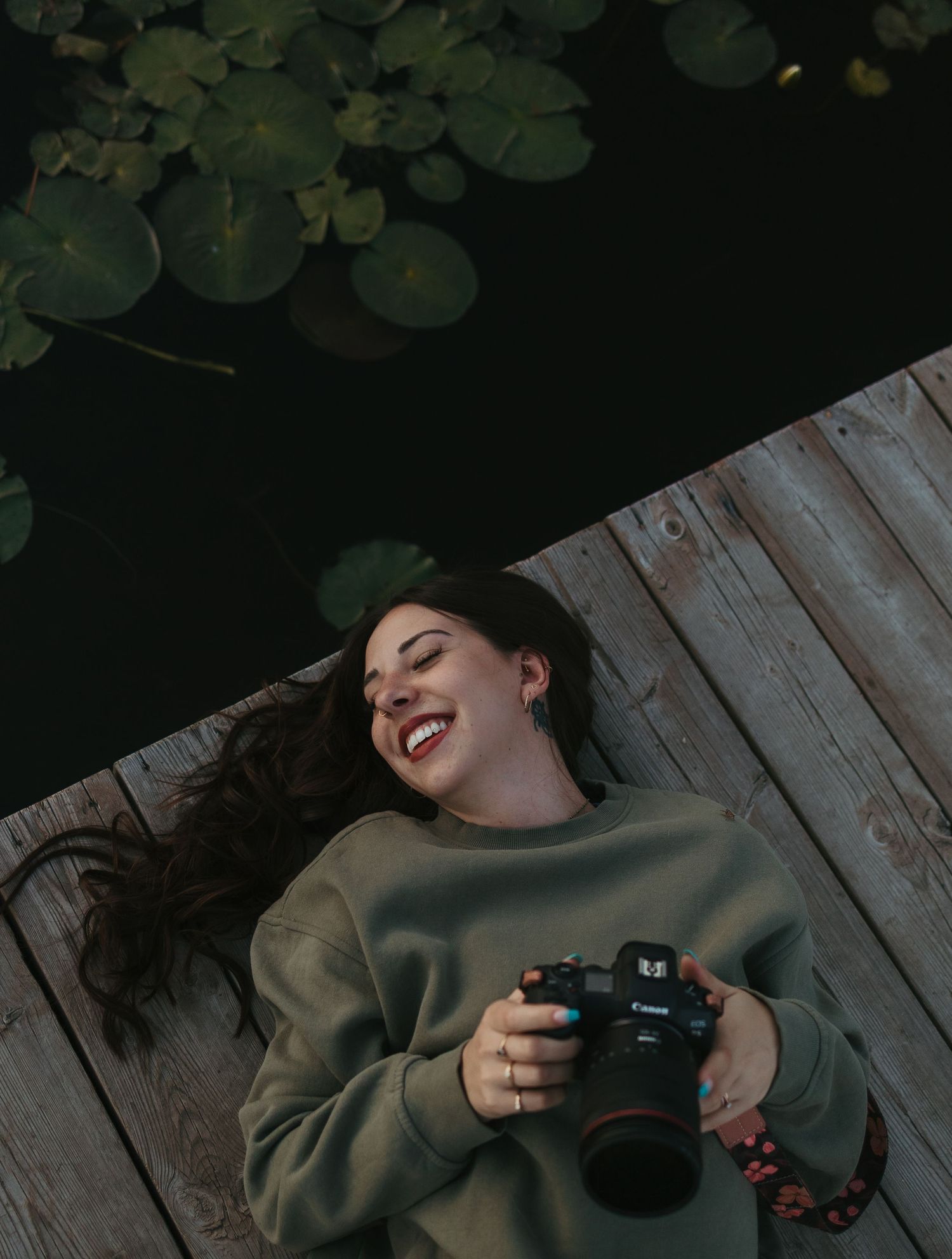 A smiling woman lying on a wooden dock holding a Canon camera, with lily pads floating on dark water behind her.