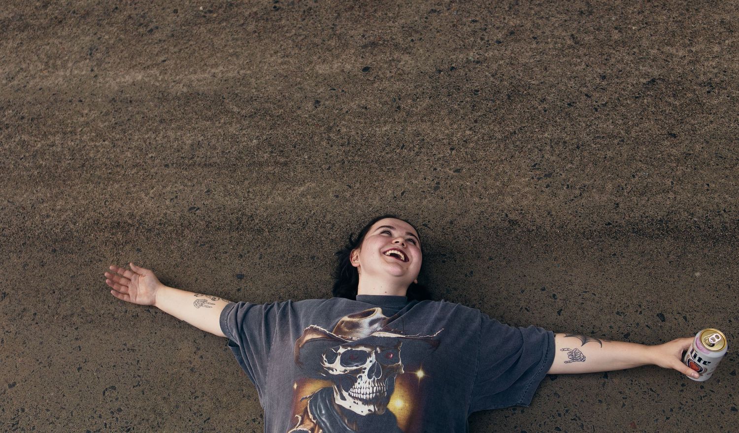 Person lying on ground laughing, wearing graphic tee, arms spread wide, holding a can.