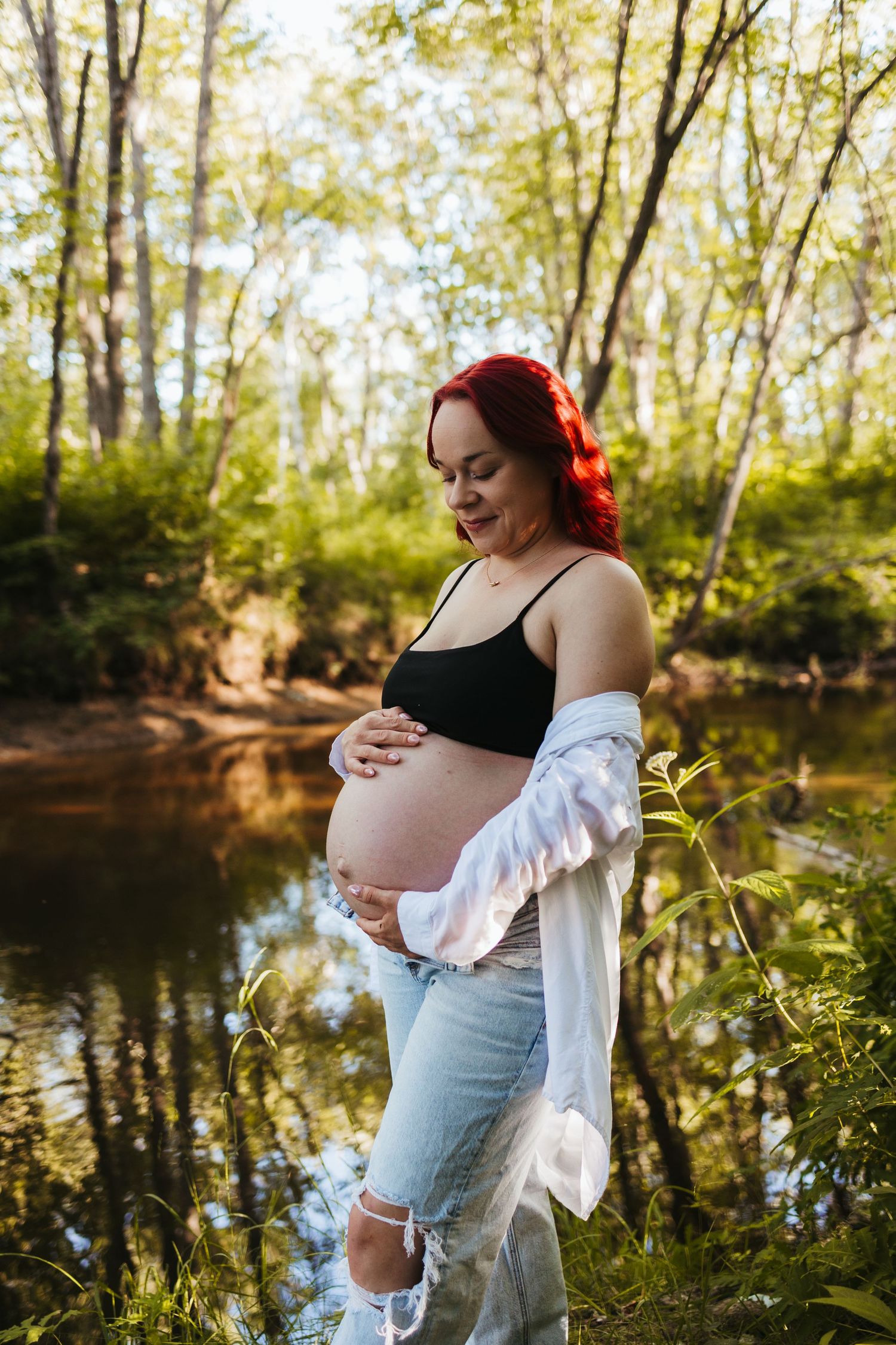 Pregnant woman with red hair in black crop top and jeans cradling baby bump outdoors near a serene forest creek.