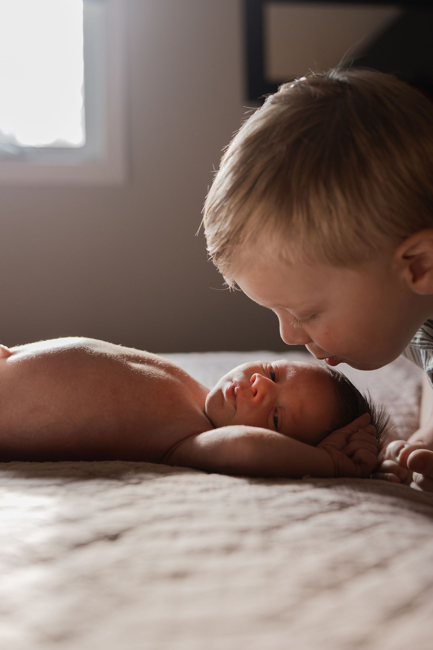 Toddler boy gently kissing newborn baby sibling lying on bed in soft natural window light.