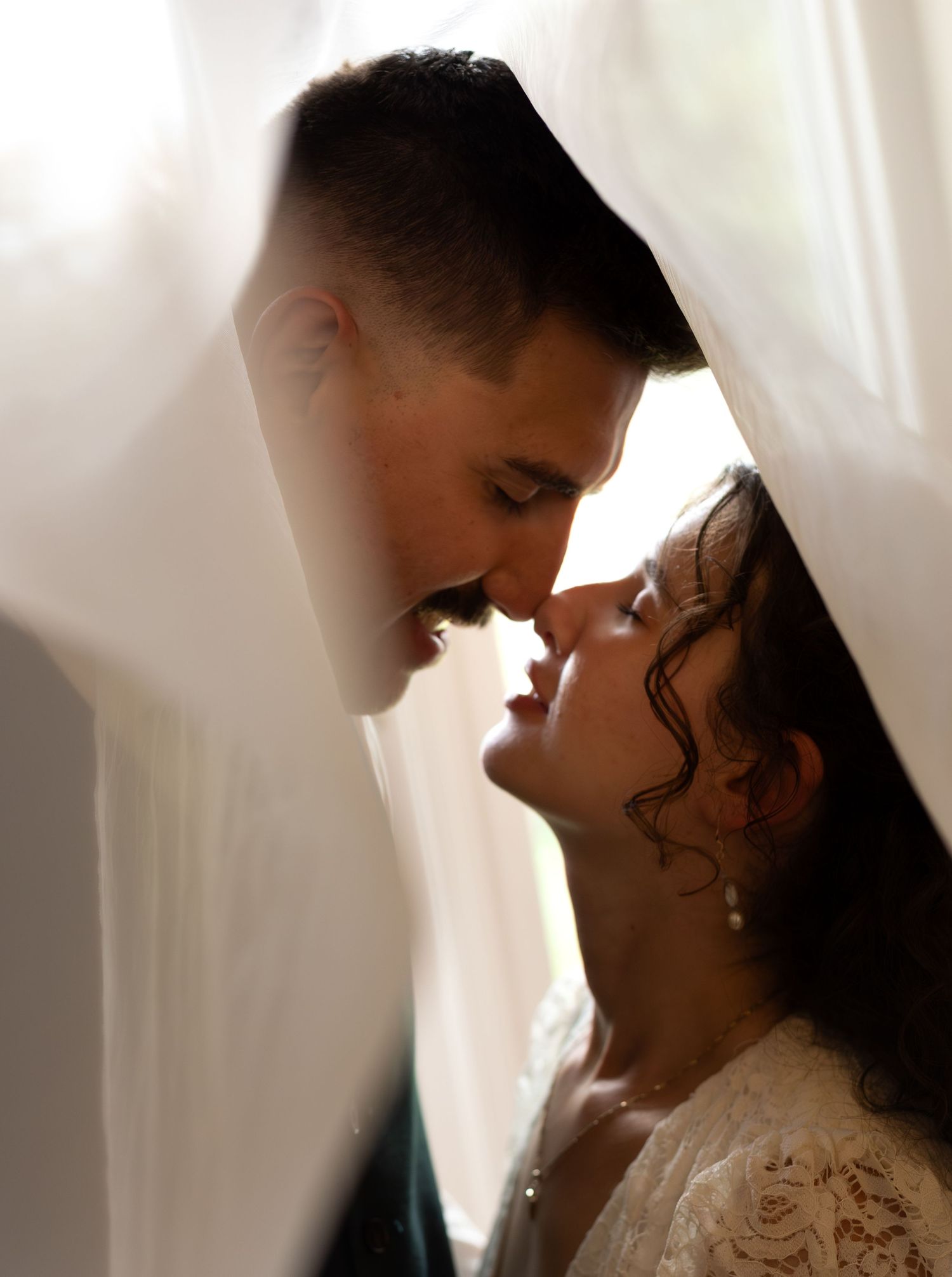 A bride and groom share a romantic moment, leaning in for a kiss framed by soft white curtains.