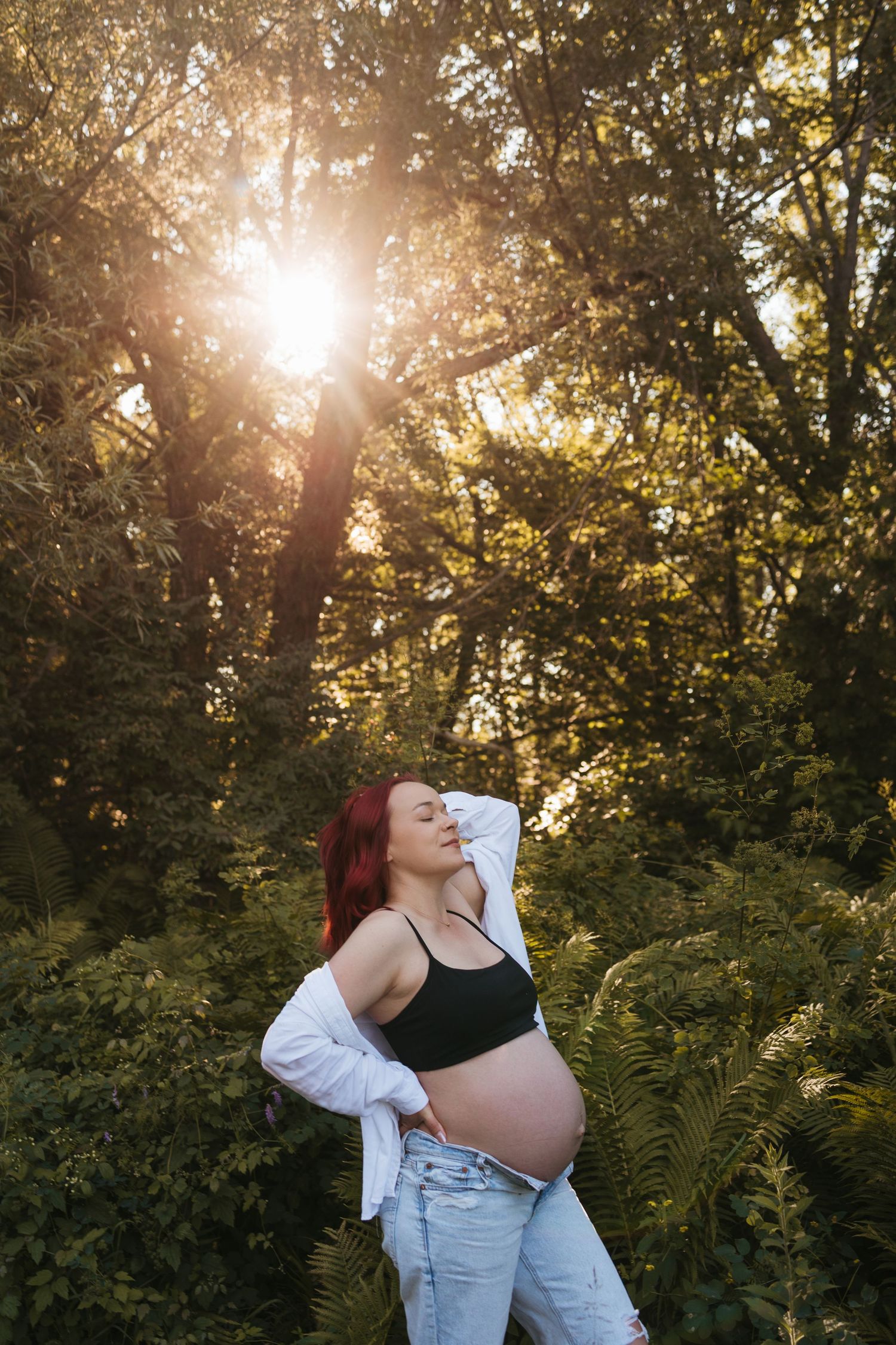 Pregnant woman with red hair poses outdoors in golden sunset light, wearing black crop top and jeans among lush green ferns.