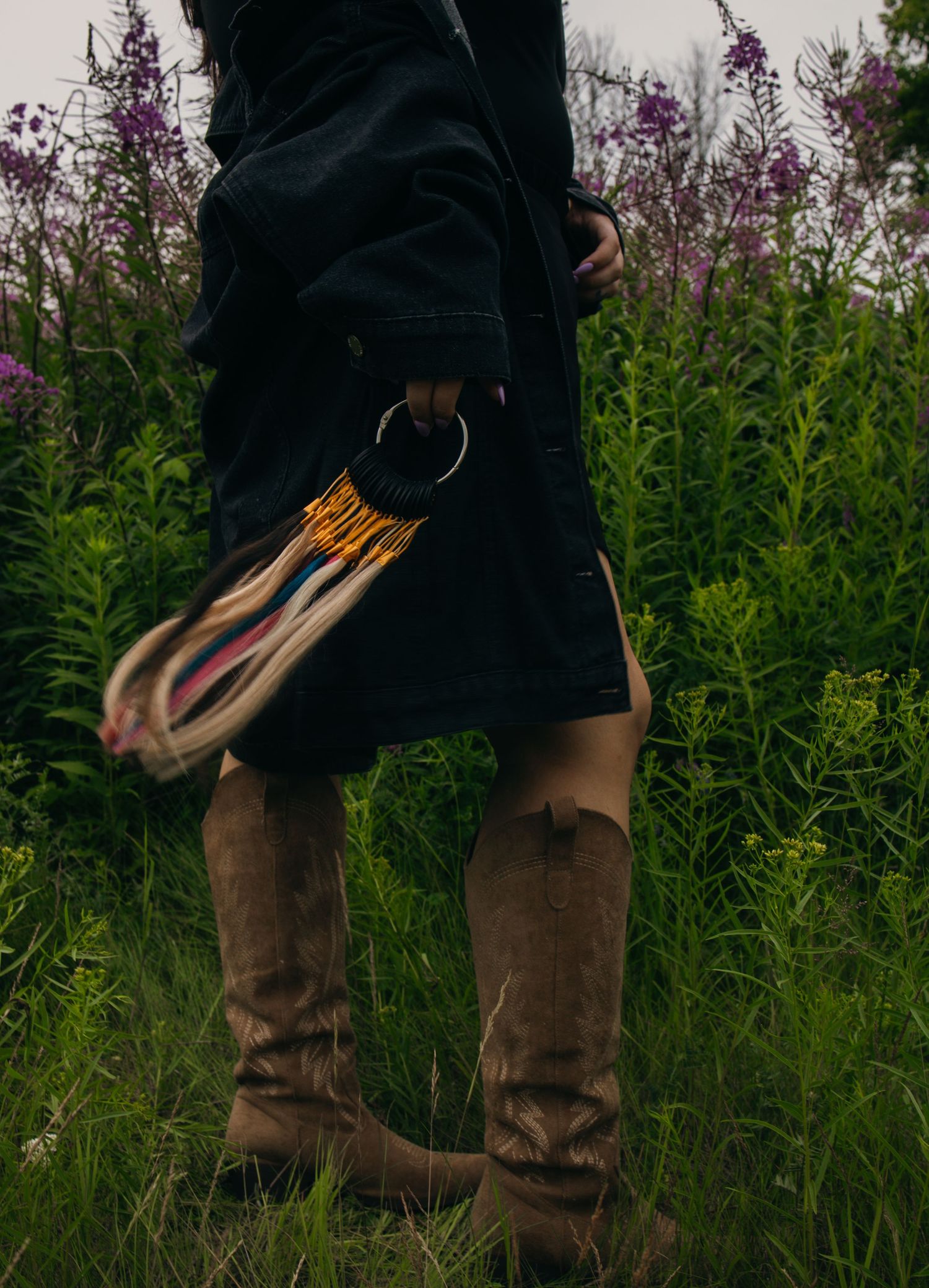 Person in black coat and tall brown boots holding colorful fringe bag, standing in lush green field with purple wildflowers.