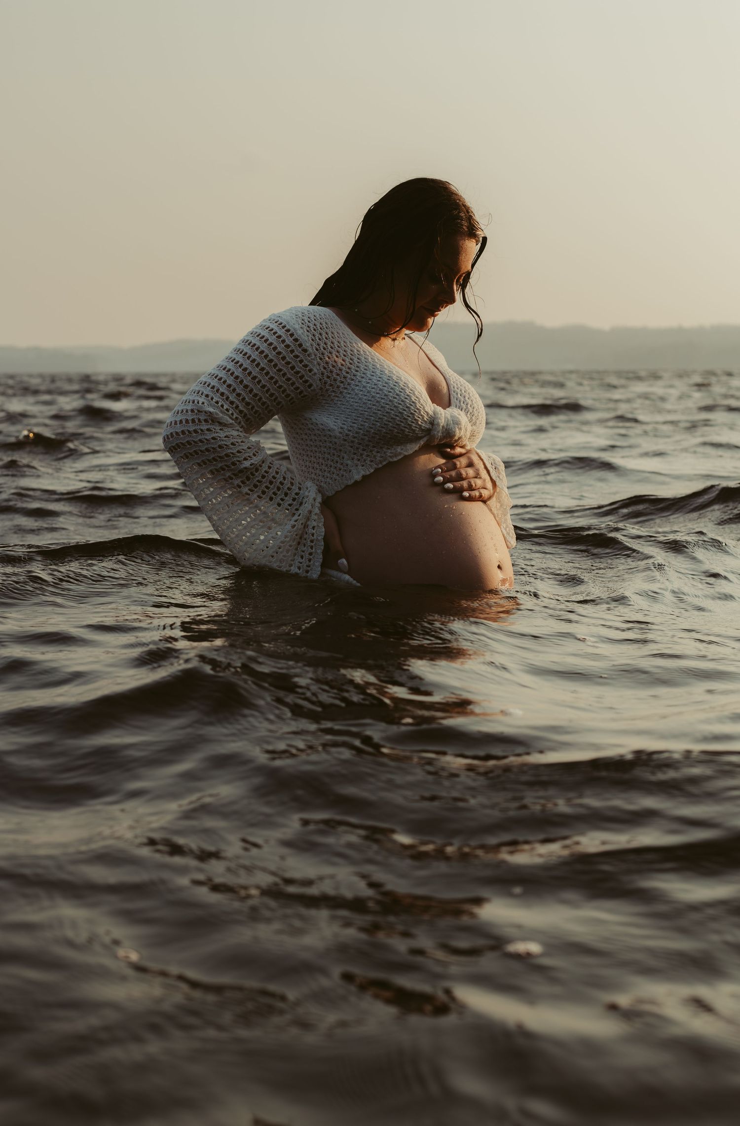 Pregnant woman in white crochet outfit standing in ocean waves at sunset, cradling her baby bump.
