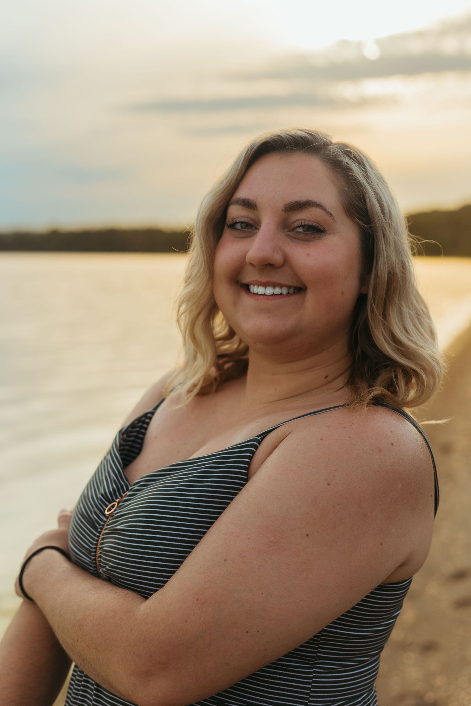 A cheerful blonde woman in a striped top stands with arms crossed on a sunny beach at golden hour.