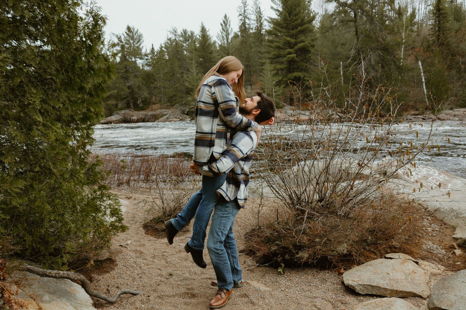 Couple sharing romantic kiss as man lifts woman near forest river.