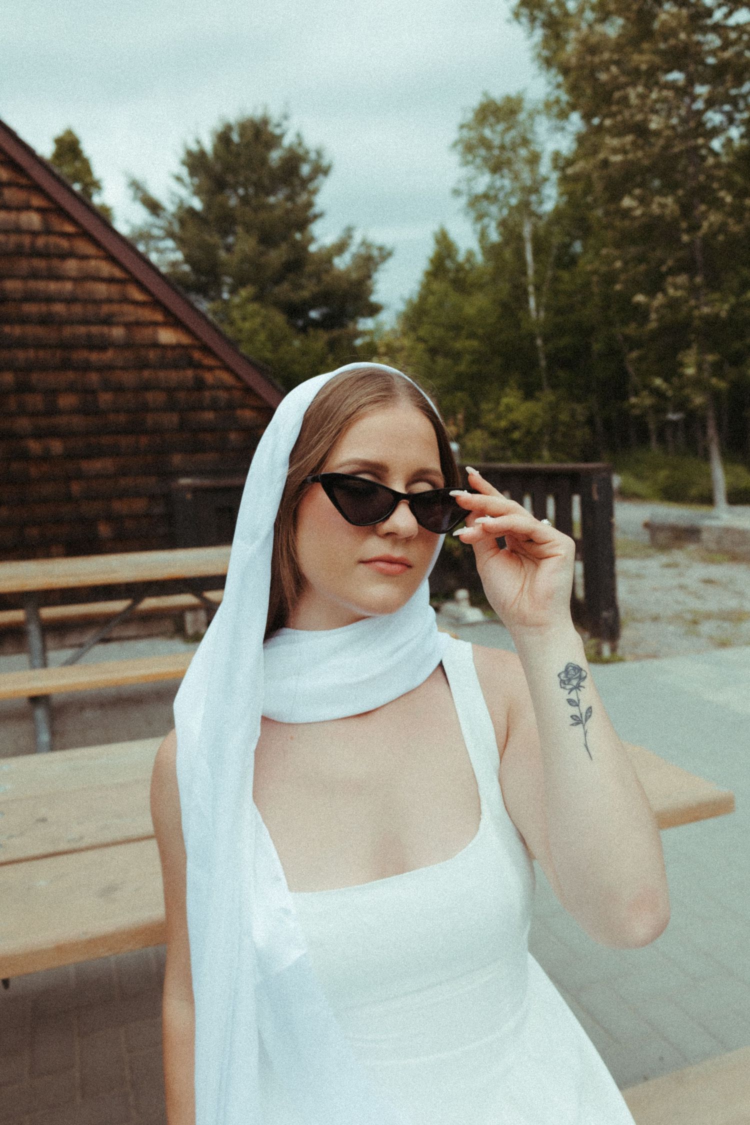 Elegant bride in white halter dress and veil posing with cat-eye sunglasses outdoors.