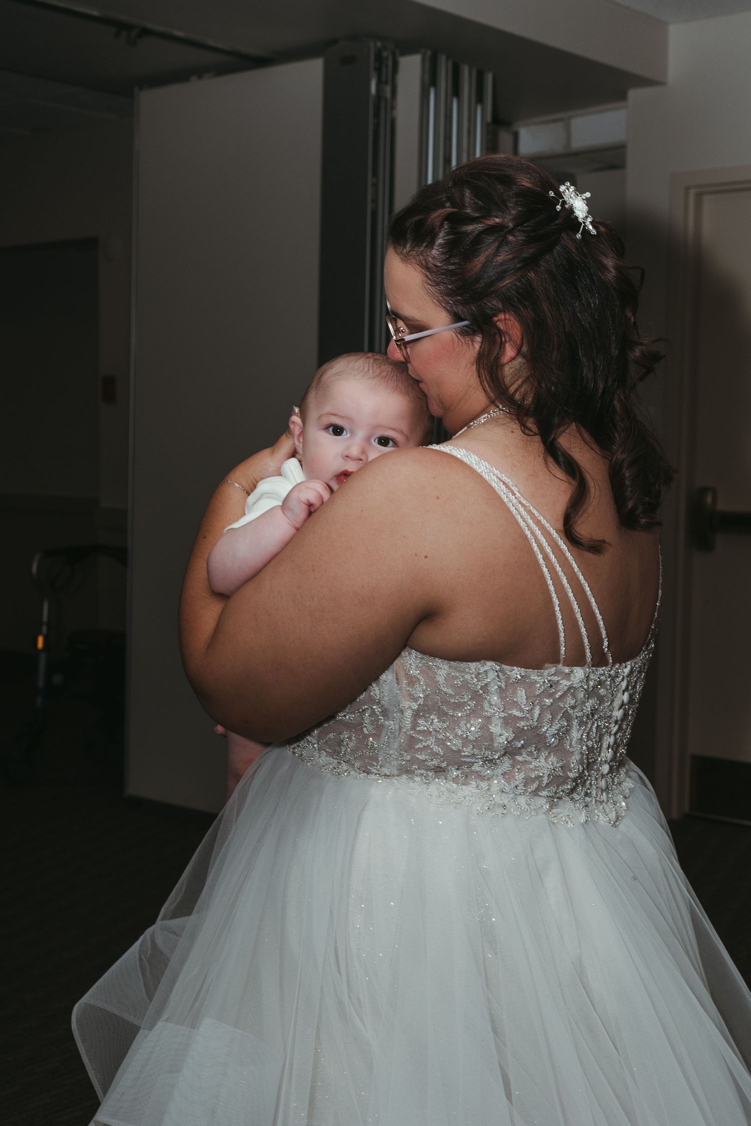 Bride in lace wedding gown with strappy back holding and kissing a newborn baby at wedding reception.
