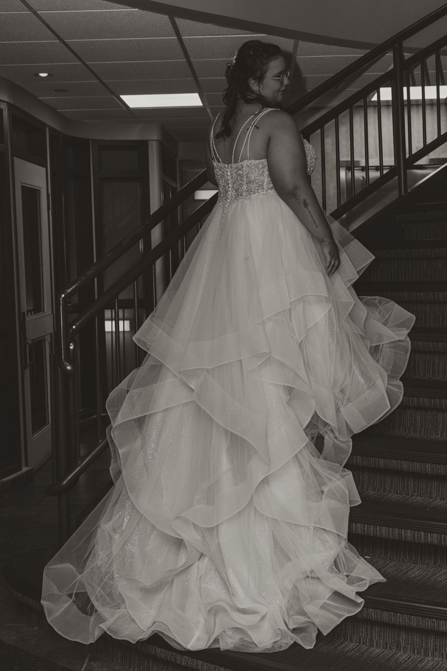A bride in a layered ballgown wedding dress stands on a staircase in black and white.