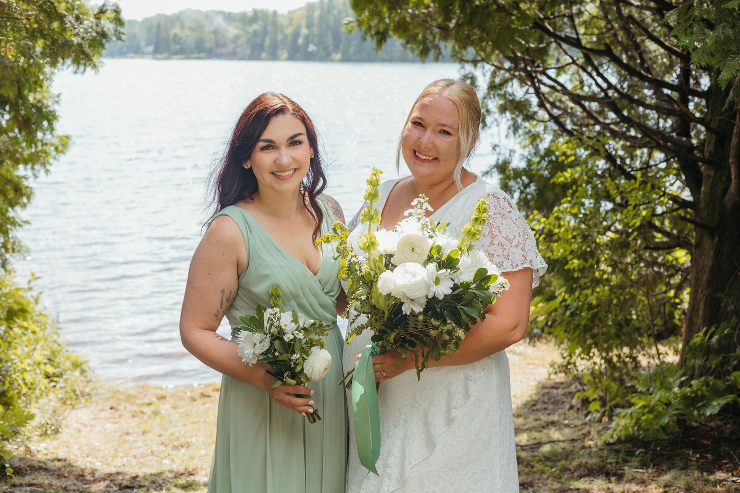 Bride in white lace dress and bridesmaid in sage green gown holding white bouquets by a lake.