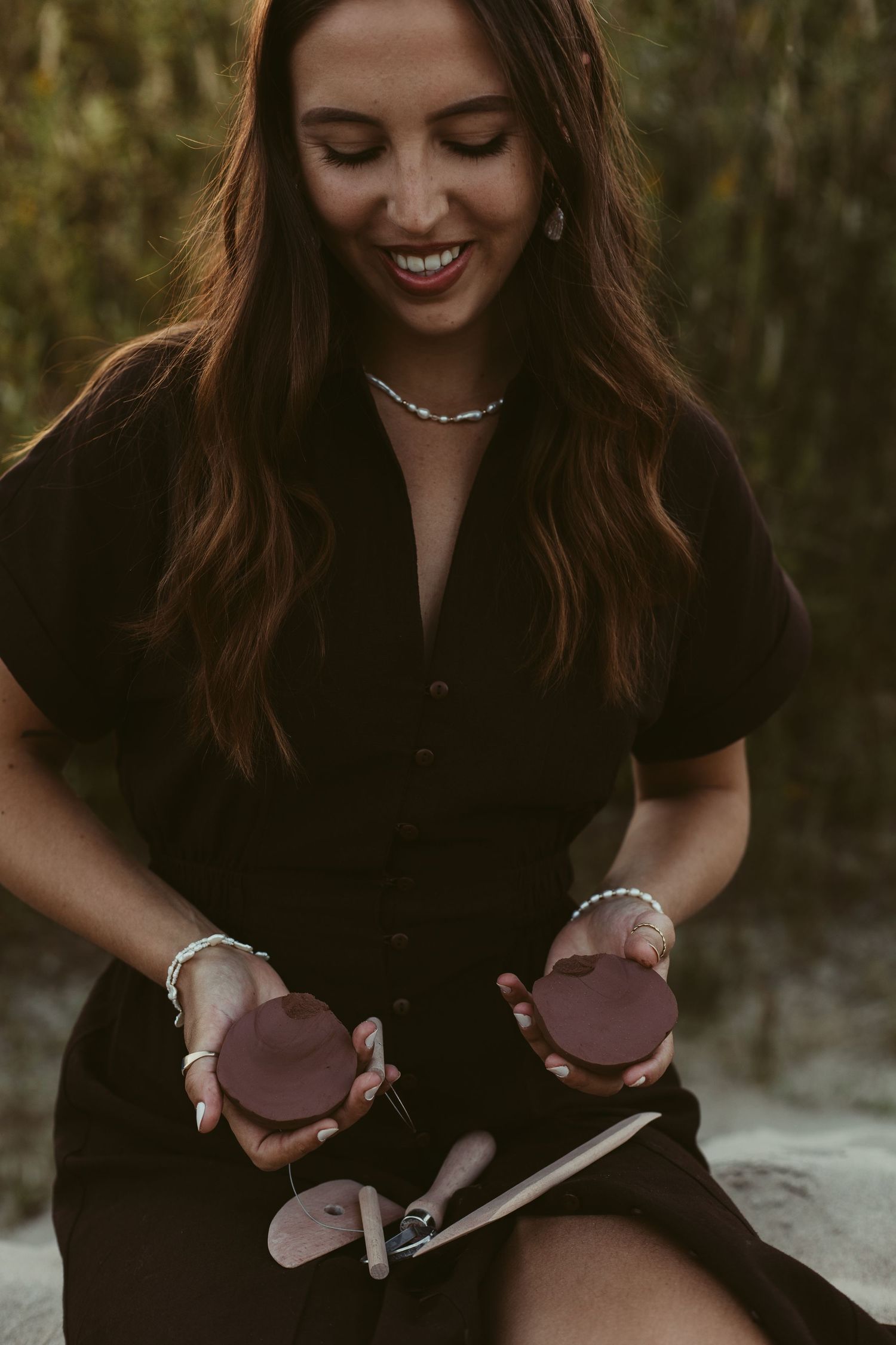 Smiling brunette woman holding two dark red ceramic discs while wearing pearl jewelry outdoors.