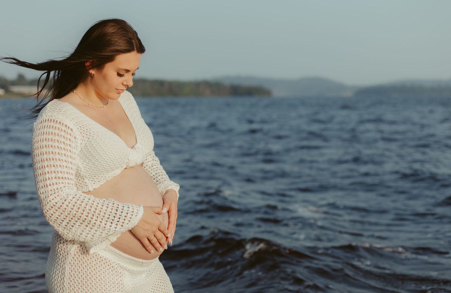 Pregnant woman in white crochet dress holding baby bump by the ocean on a sunny day.