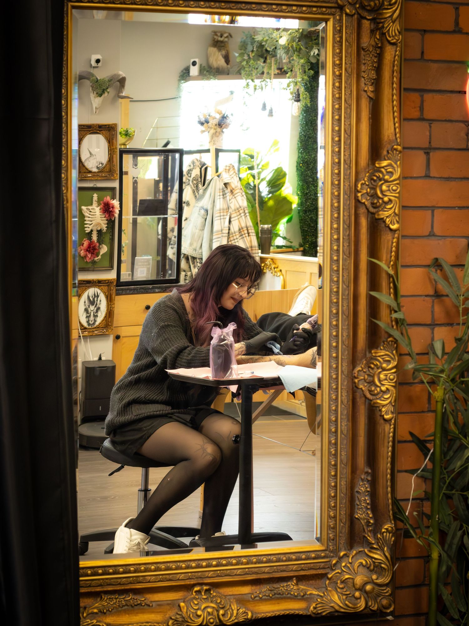 Woman reflected in ornate gold mirror working at small table inside a cozy decorated studio.
