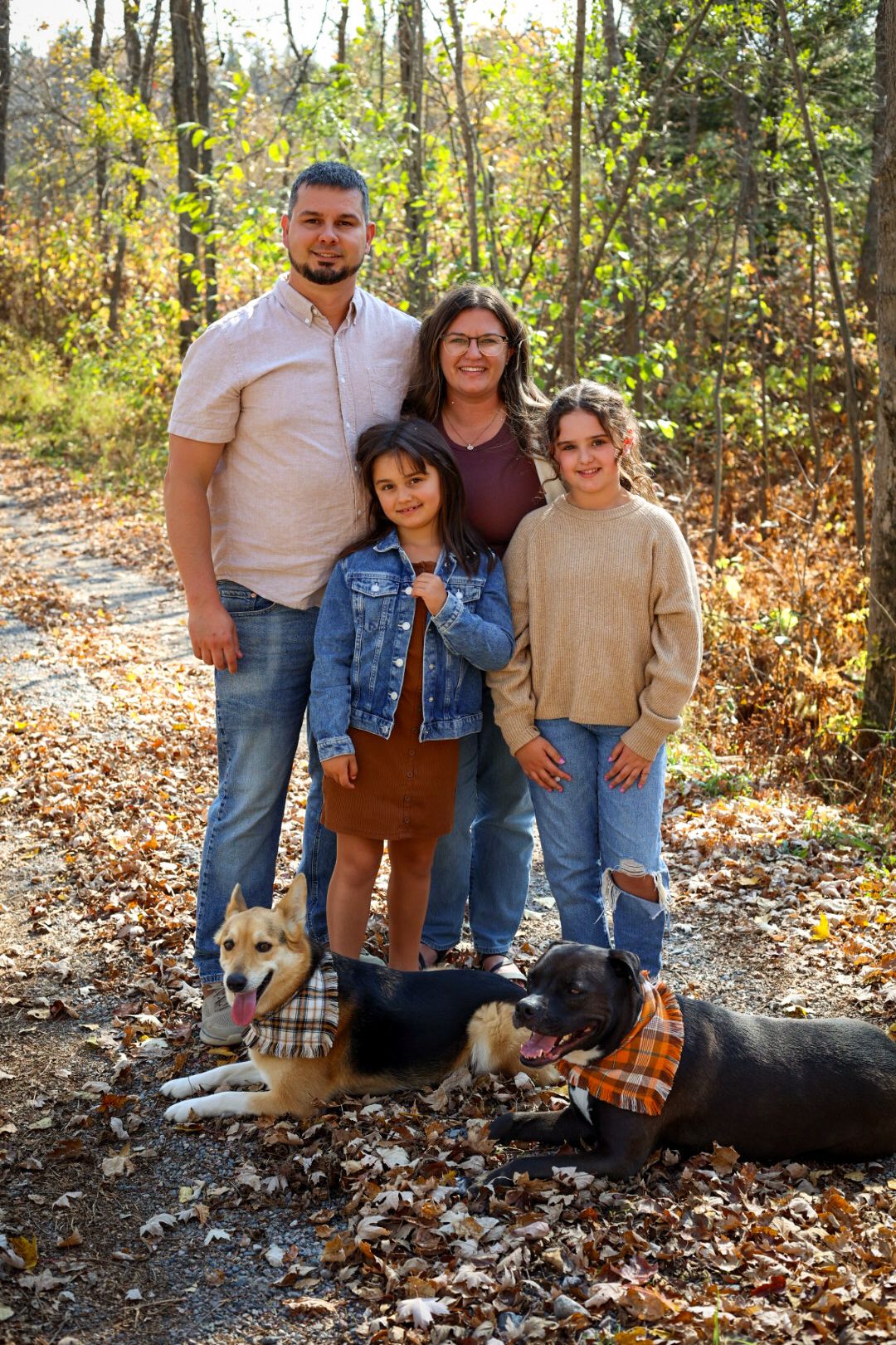 Family of four poses with two dogs wearing plaid bandanas among autumn leaves in a wooded setting.