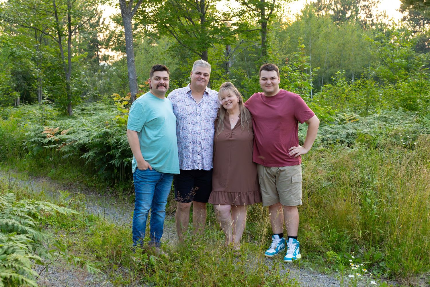 Family of four smiles together on wooded path during golden summer sunset.